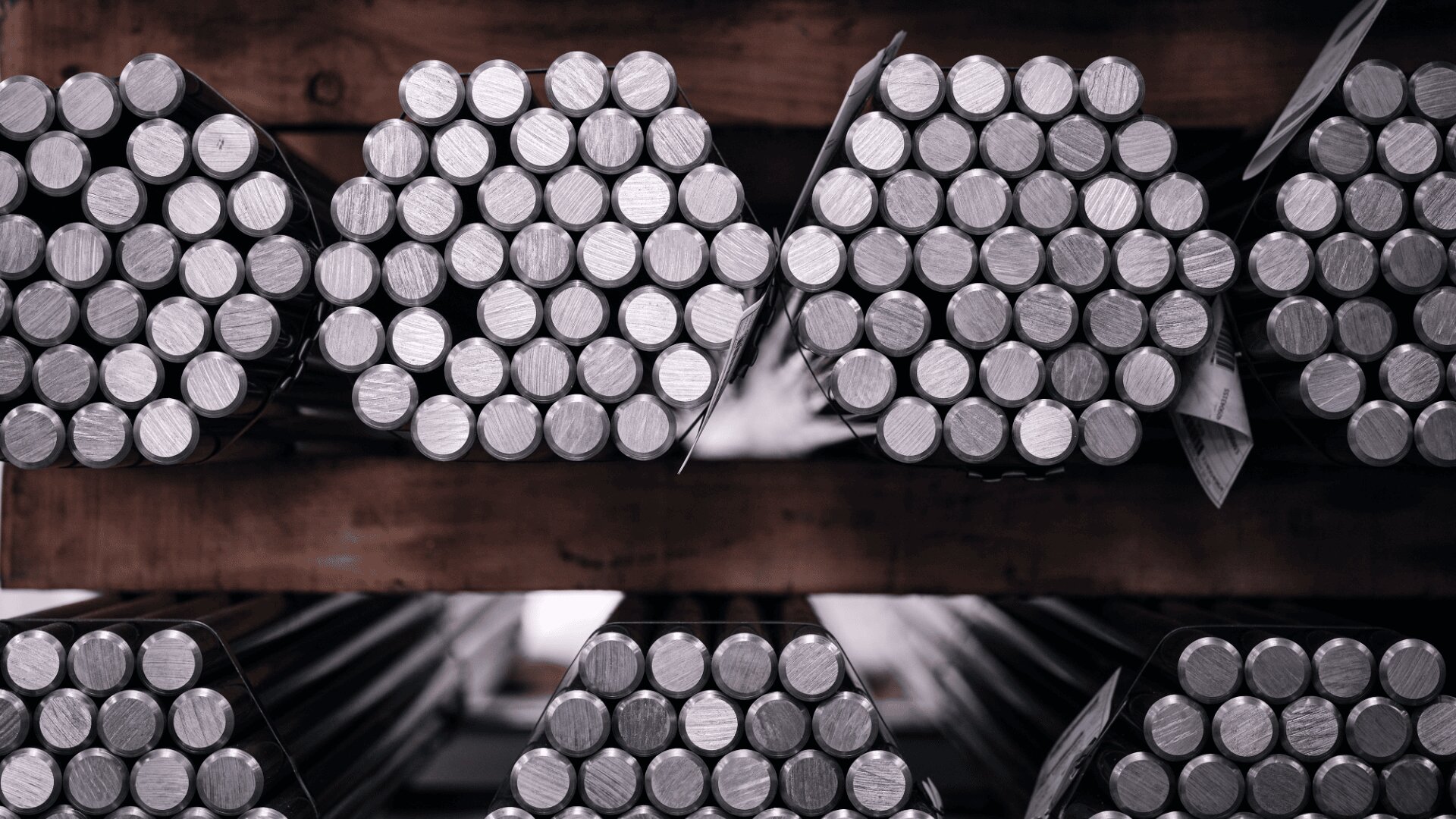 Stapel von Metallstangen sind in Bündeln angeordnet und auf Holzregalen gelagert. Von einem Ende aus betrachtet, sind die runden Enden der Stangen in einer industriellen Umgebung zu sehen.