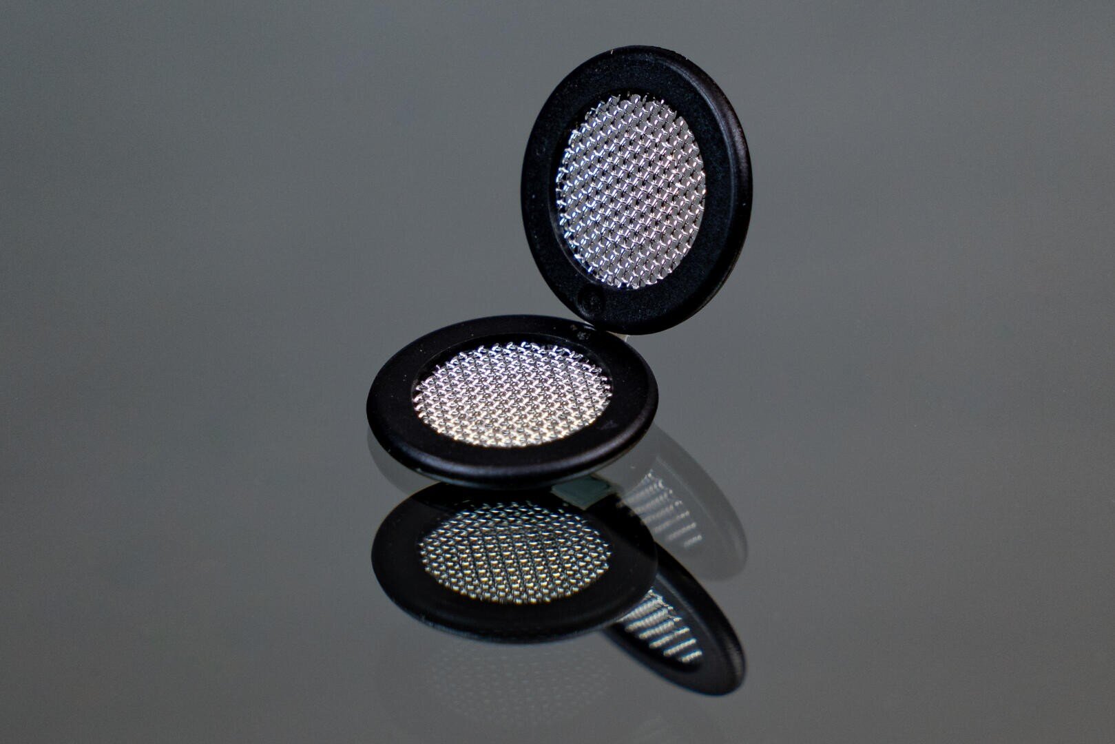 A small, round, black compact with a mesh-like surface is open and standing upright on a reflective gray surface, showing both the lid and the base’s textured interior.