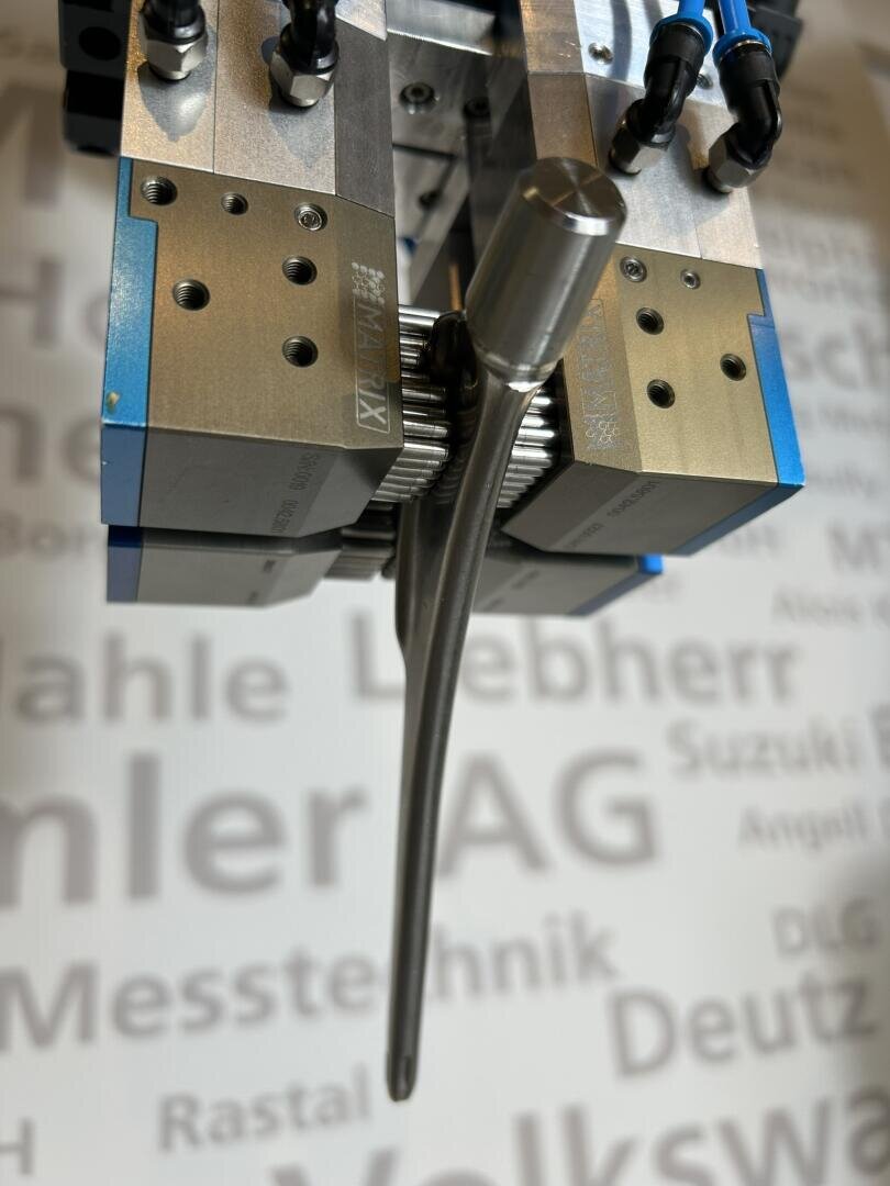 Close-up of a robot gripper holding a metal automobile valve. In the background is a blurred wall with the names of various car manufacturers, including Volkswagen, MAN and Deutz.