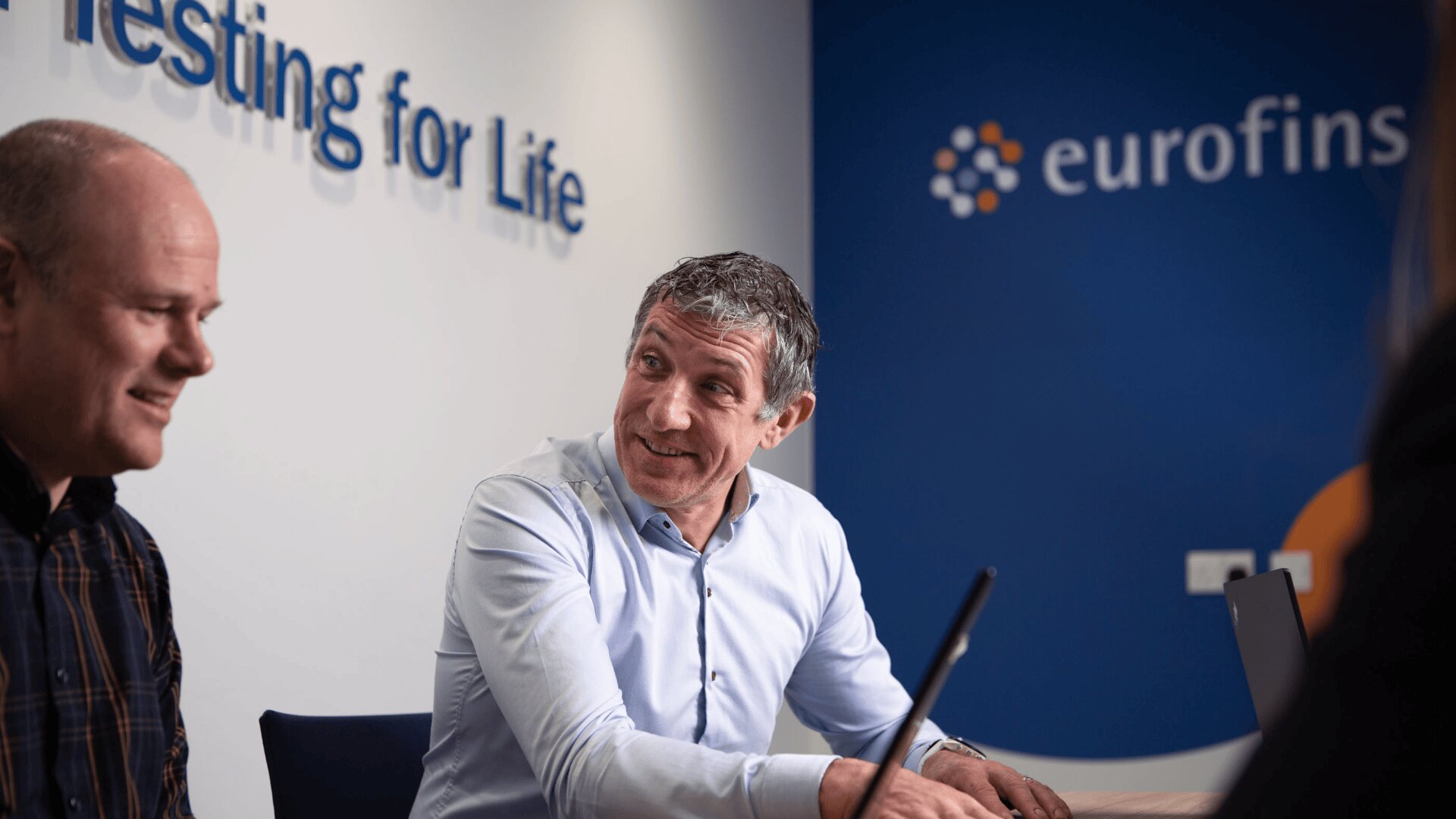Zwei Männer sitzen mit Laptops an einem Tisch in einem Besprechungsraum. An der Wand hinter ihnen sind die Worte „Testing for Life“ und das Eurofins-Logo zu sehen. Ein Mann lächelt und blickt zum anderen.