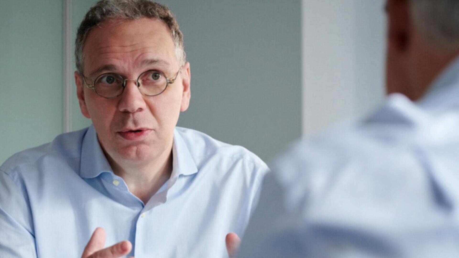 A man with short, gray hair and glasses, wearing a light blue shirt, sits indoors and gestures while speaking to another person whose back is to the camera.