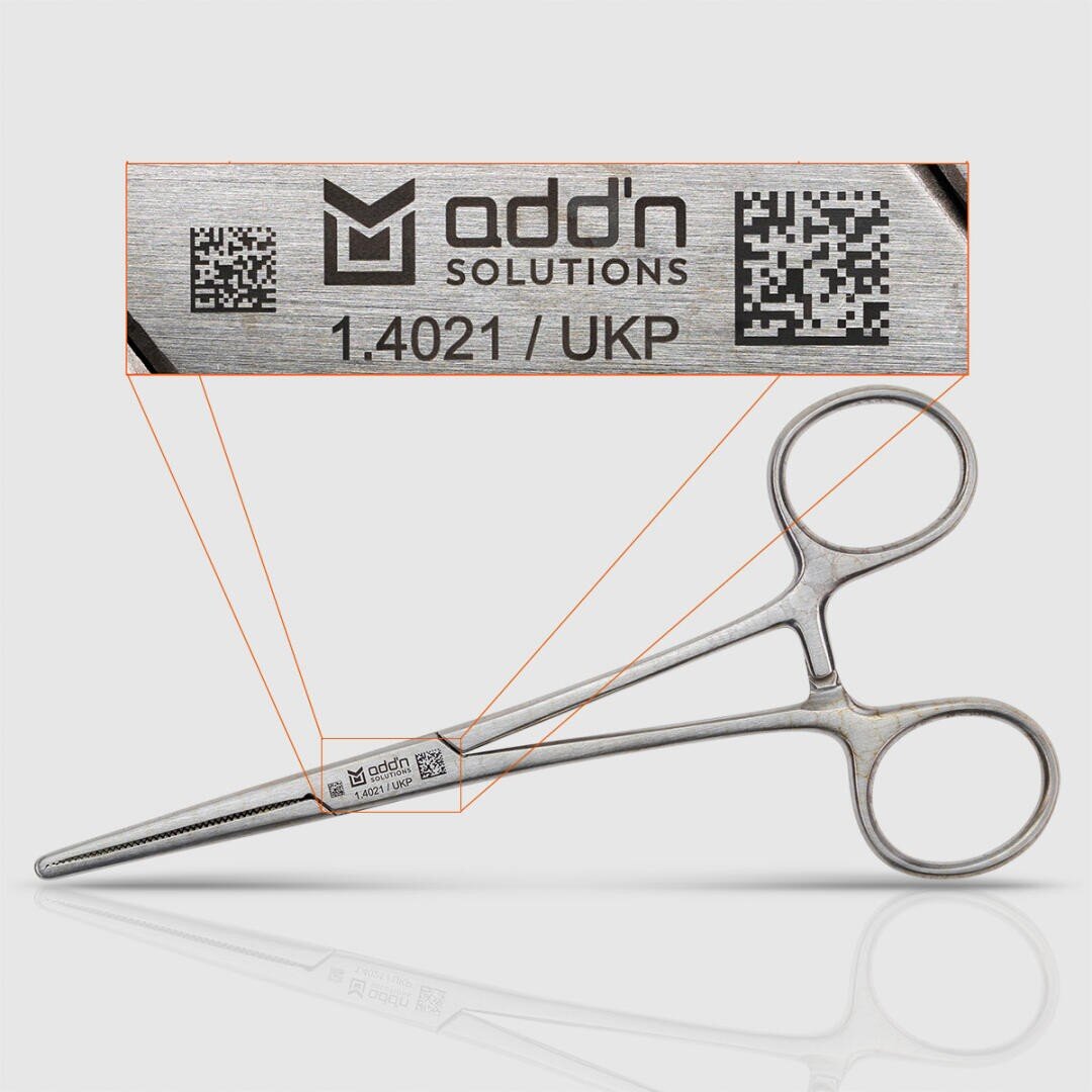 A surgical metal instrument with finger rings and a locking mechanism, shown with a zoom of the engraved label with the inscription addn SOLUTIONS, 1.4021 / UKP and a QR code.