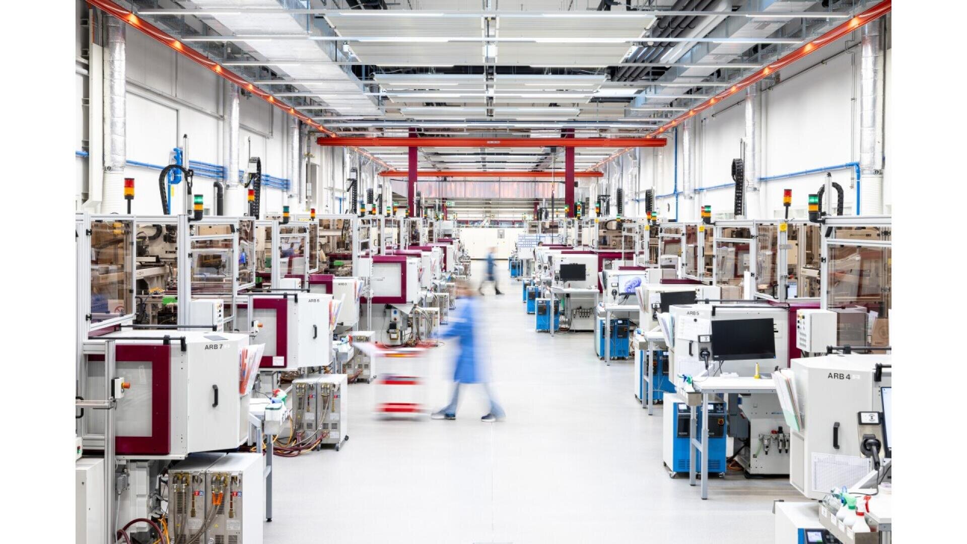 Eine geräumige, hell erleuchtete Fabrikhalle mit Reihen automatisierter Maschinen und Computerbildschirmen. Eine Person in blauer Arbeitskleidung bewegt sich schnell durch den Mittelgang und wirkt durch die Bewegung verschwommen.