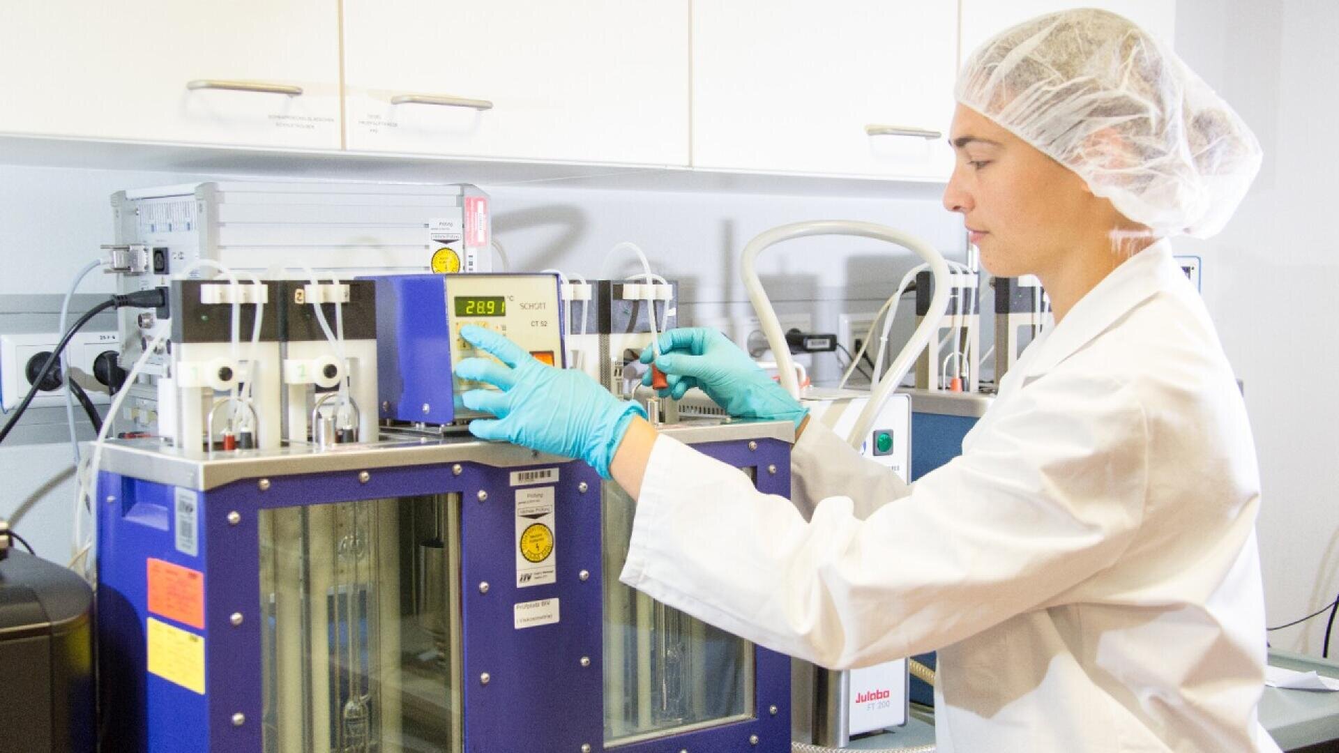 A person in a lab coat, hairnet, and gloves operates scientific equipment in a laboratory setting, adjusting controls on a blue and silver machine with digital displays and tubes.