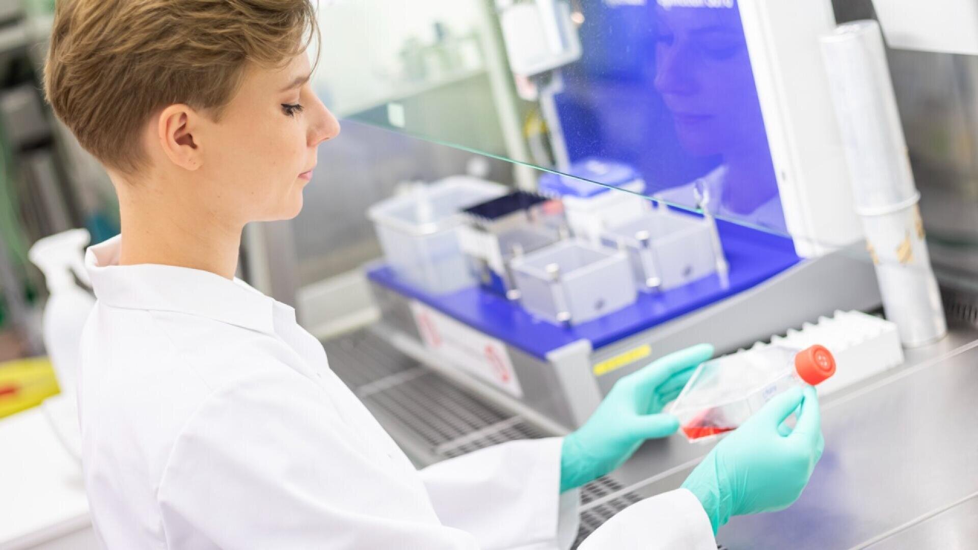 A scientist in a white lab coat and green gloves examines a container with red liquid inside a laboratory, working next to scientific equipment and a protective glass barrier.