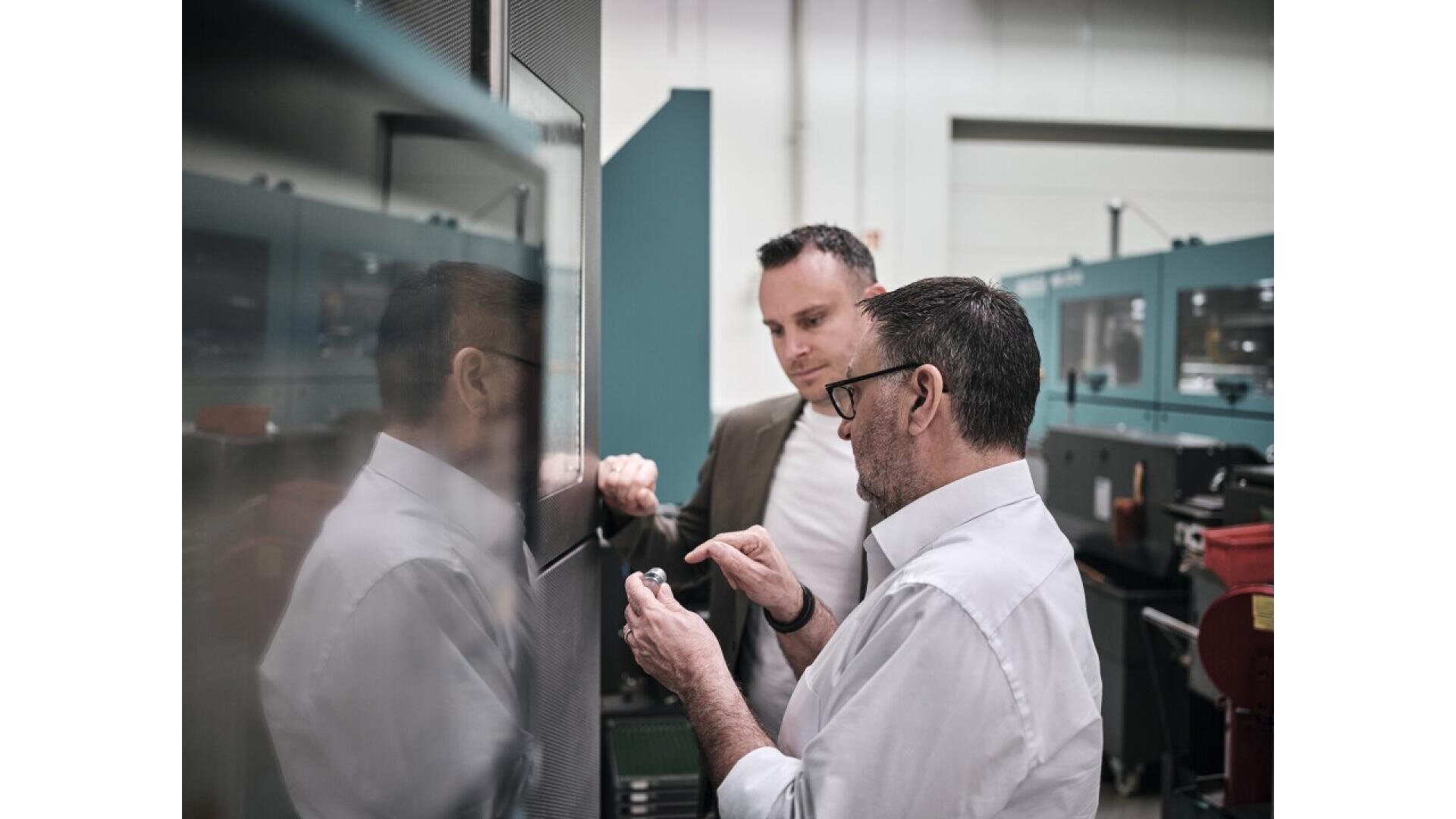 Two men in business attire stand near industrial equipment in a factory setting. One man is explaining or showing something small, possibly a part or component, to the other man. The surroundings are clean and modern.