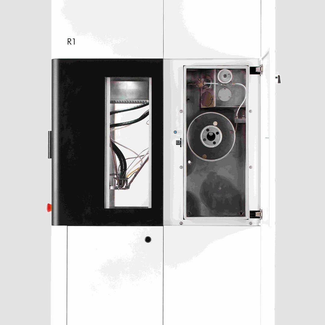 Eine weiße Industriemaschine mit einer schwarzen Blende auf der linken Seite, beschriftet mit R1. Die rechte Seite hat eine transparente Tür, durch die man mechanische Komponenten wie Drähte, Riemenscheiben und Zahnräder im Inneren sehen kann.