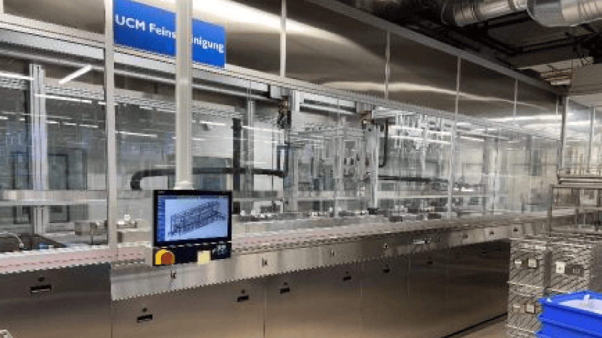 Eine moderne Industriereinigungsmaschine aus rostfreiem Stahl steht in einer Fabrikhalle. Auf einem Kontrollmonitor ist ein Diagramm zu sehen, darüber ein blaues Schild mit der Aufschrift UCM Feinstreinigung. Hinter Glasscheiben sind verschiedene Rohre und Geräte zu sehen.