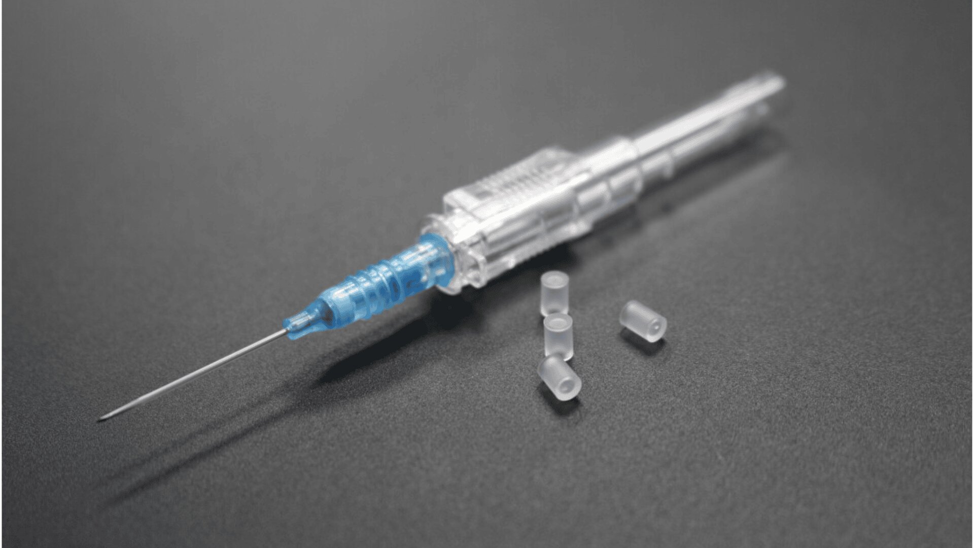 A close-up of a medical syringe with a blue hub and clear protective cap, resting on a dark surface beside three small clear plastic caps.
