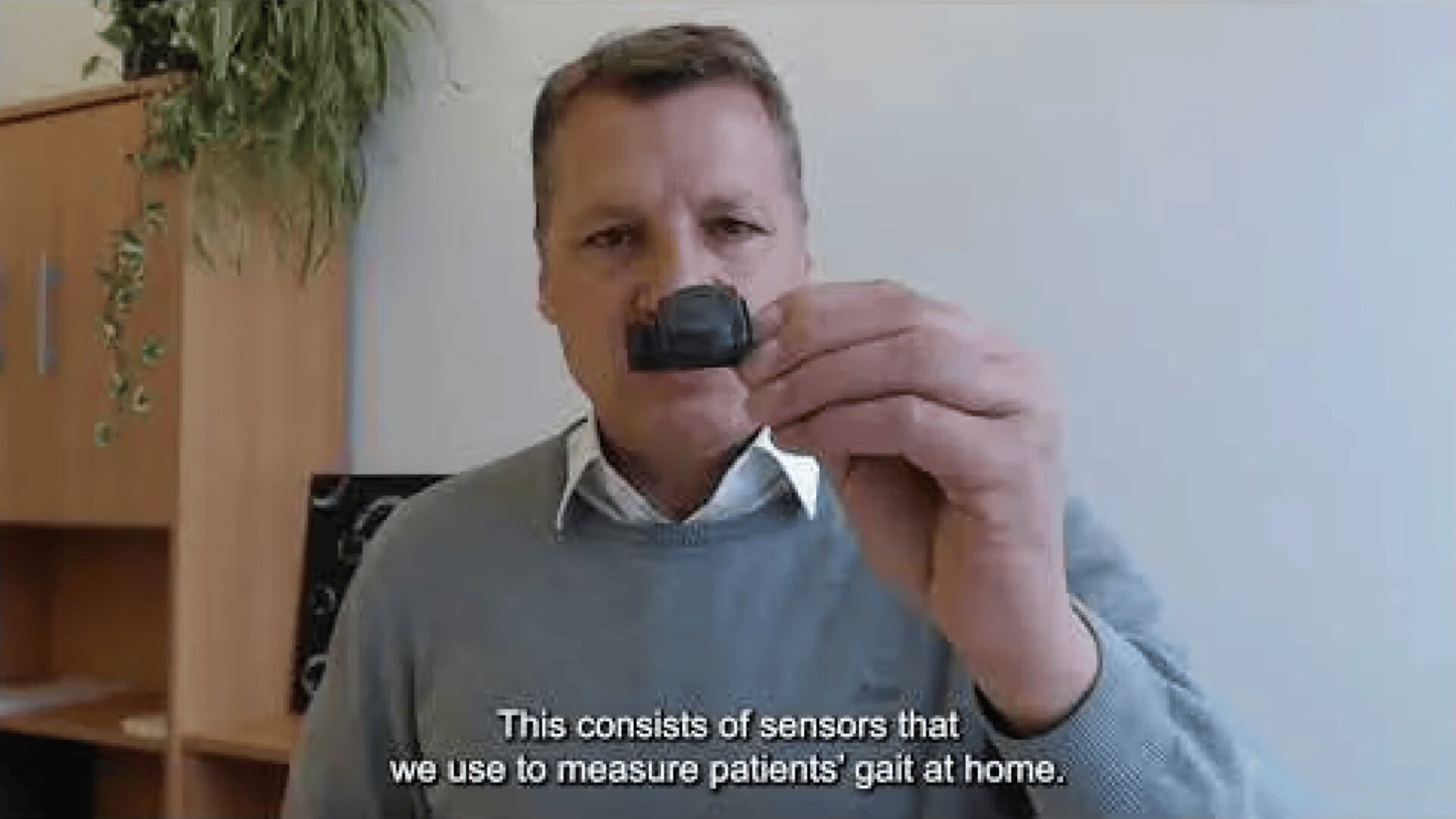 A man in a light blue sweater holds a small black sensor up to the camera. The caption reads: These are sensors we use to measure patients gait at home.