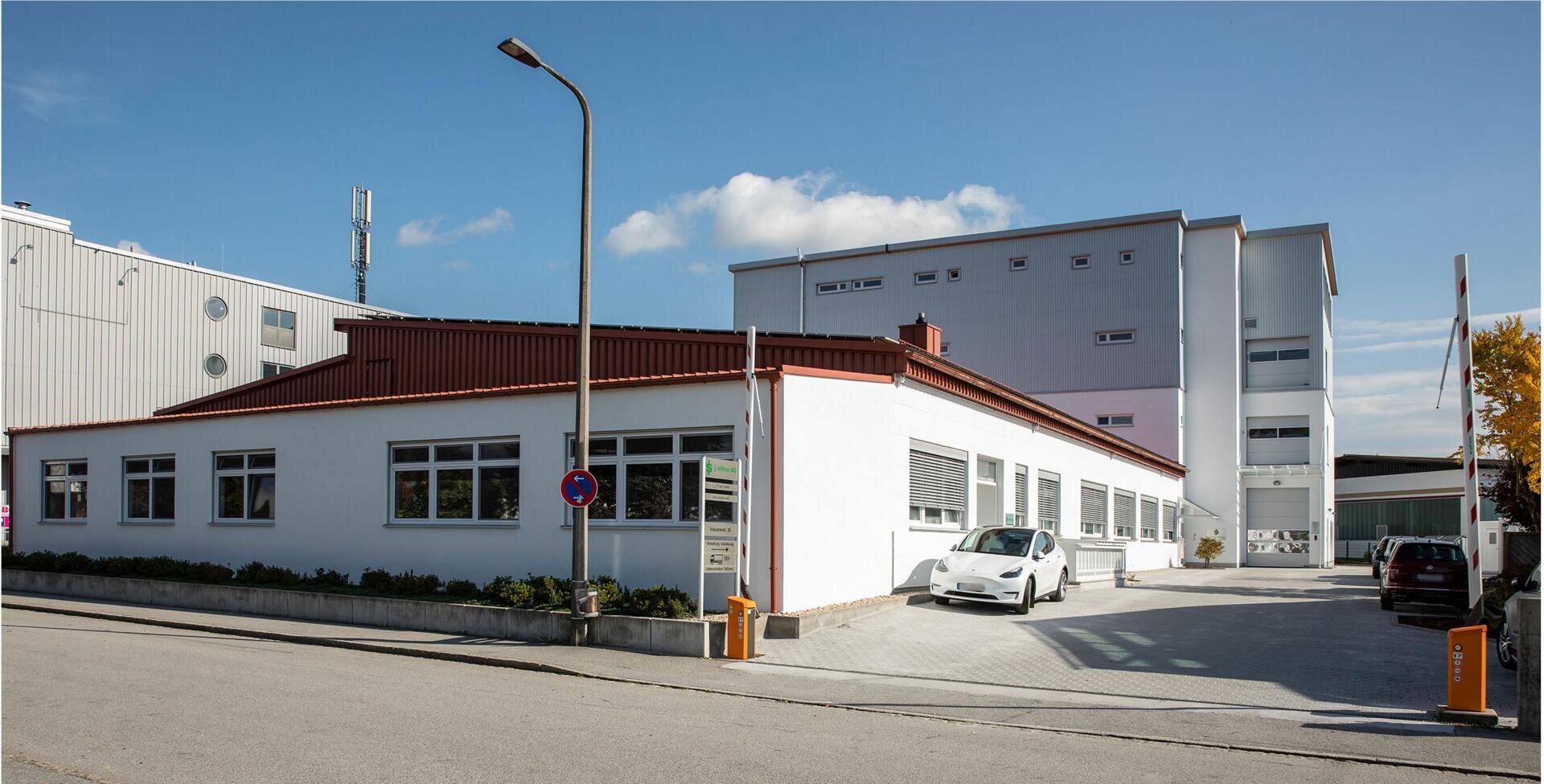 Ein modernes Industriegebäude mit weißen Wänden und roten Zierleisten sowie großen Fenstern. Ein weißes Auto parkt davor, ein weiteres steht in der Nähe des Eingangs. Der Himmel ist klar, nur wenige Wolken ziehen sich durch.