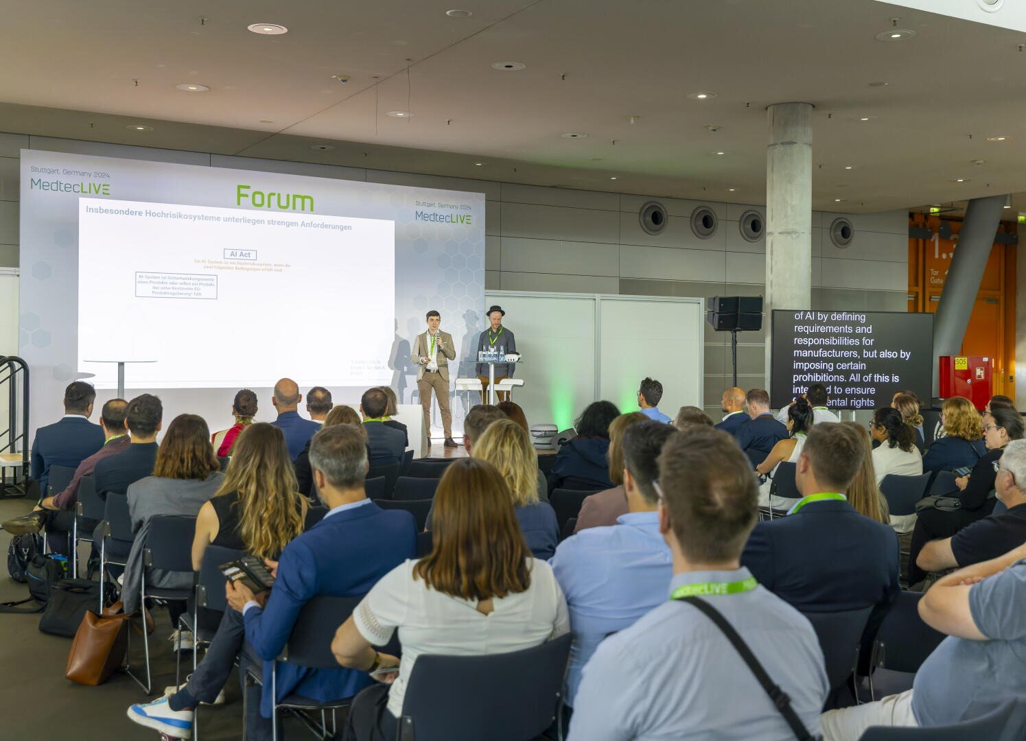 Eine Gruppe von Personen sitzt vor einer Bühne, auf der zwei Redner bei einem Fachforum eine Folie präsentieren. Das Publikum hört aufmerksam in einem modernen Konferenzraum zu, in dem eine große Leinwand und Untertitel zu sehen sind.