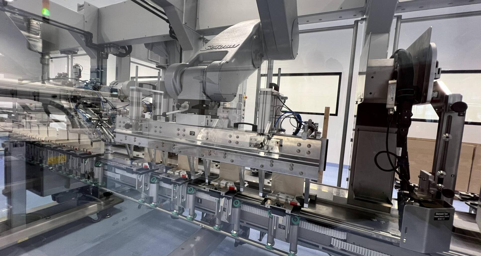 Eine moderne automatisierte Maschine in einer sterilen Fabrikumgebung füllt Fläschchen oder Flaschen auf einem Förderband ab, wobei verschiedene mechanische Arme und Komponenten hinter Glasscheiben sichtbar sind.