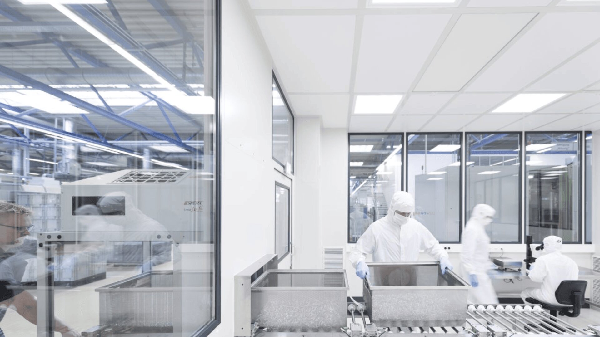 Two people in white cleanroom suits work with metal containers in a bright, modern lab with large windows. Another suited person is seen in the background at a desk. The environment appears sterile and high-tech.