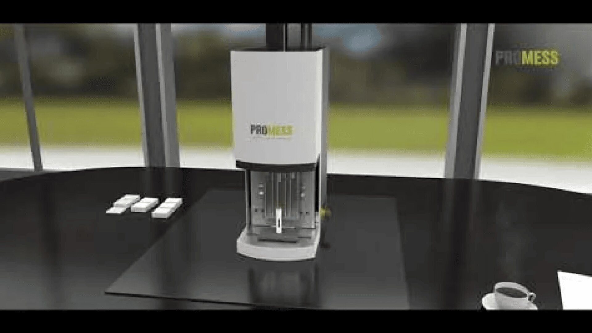 A Promess machine sits on a black table near a window, with a few white objects on the left and a cup and saucer on the right. The background shows blurred greenery outside.