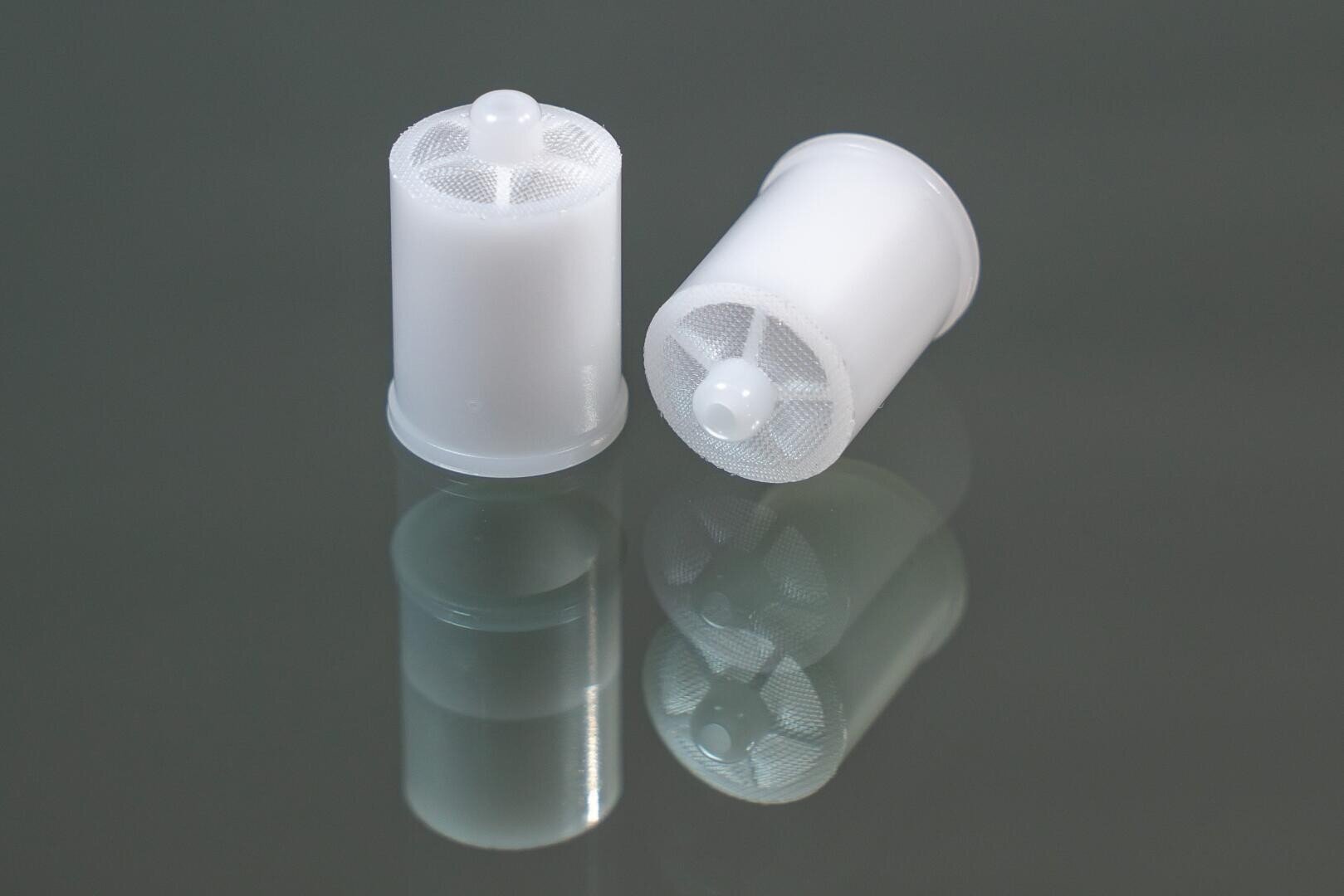 Auf einer spiegelnden grauen Fläche sind zwei kleine, zylindrische Filter aus weißem Kunststoffgewebe abgebildet; ein Filter steht aufrecht, der andere liegt auf der Seite.