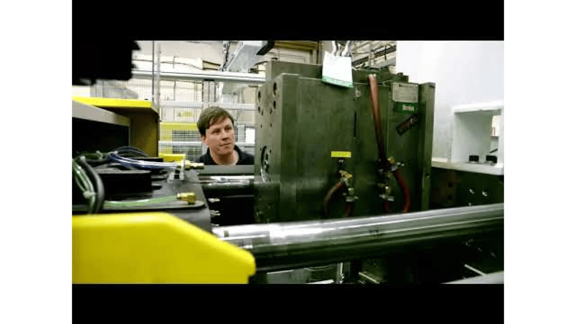 A person stands in a factory, observing industrial machinery with metal components, pipes, and cables. The setting appears to be a manufacturing or assembly area.
