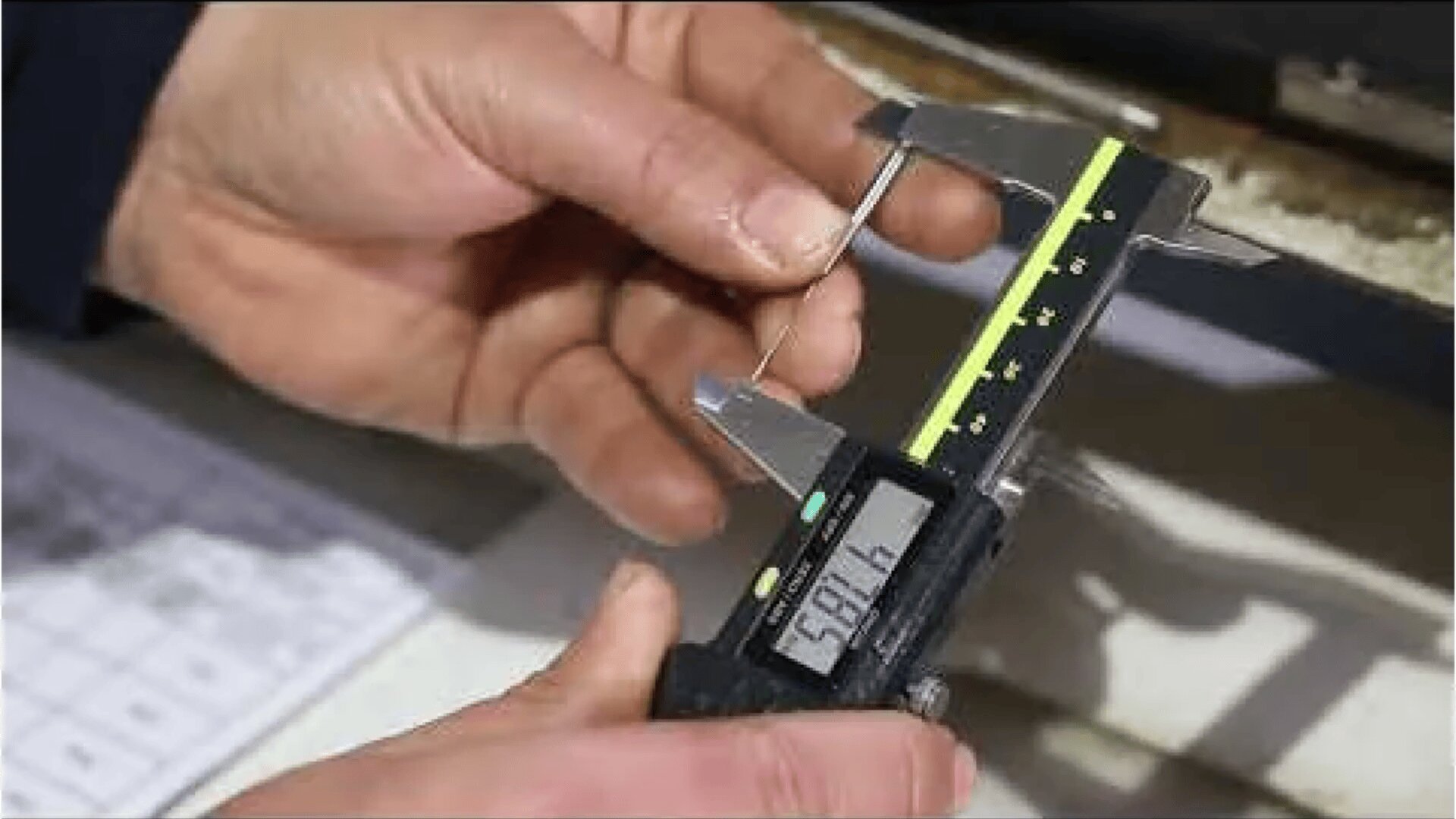 A person measures a thin metal object with a digital caliper. The calipers digital display shows the measurement value 59.16. A sheet of text is visible in the background.