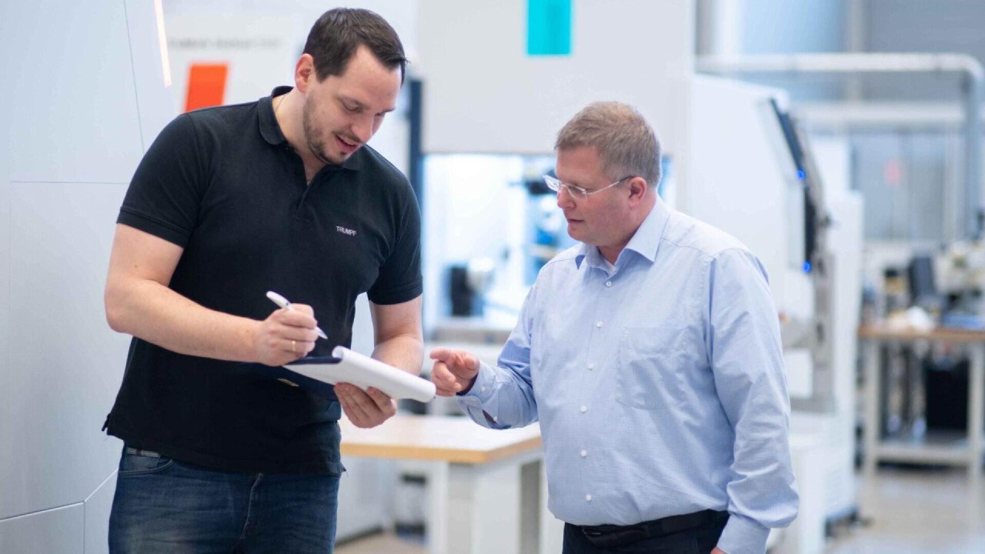 Two men standing in a modern office or lab, one in a black shirt holding a notepad and pen, the other in a light blue shirt, both engaged in conversation and looking at the notepad.