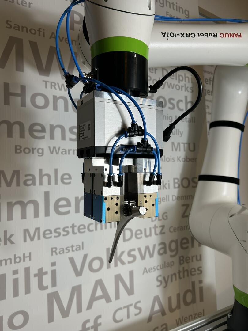 Close-up of a robotic arm with a mechanical gripper and blue hoses standing in front of a wall on which various names of automotive and industrial companies are displayed in different sizes and shades of gray.
