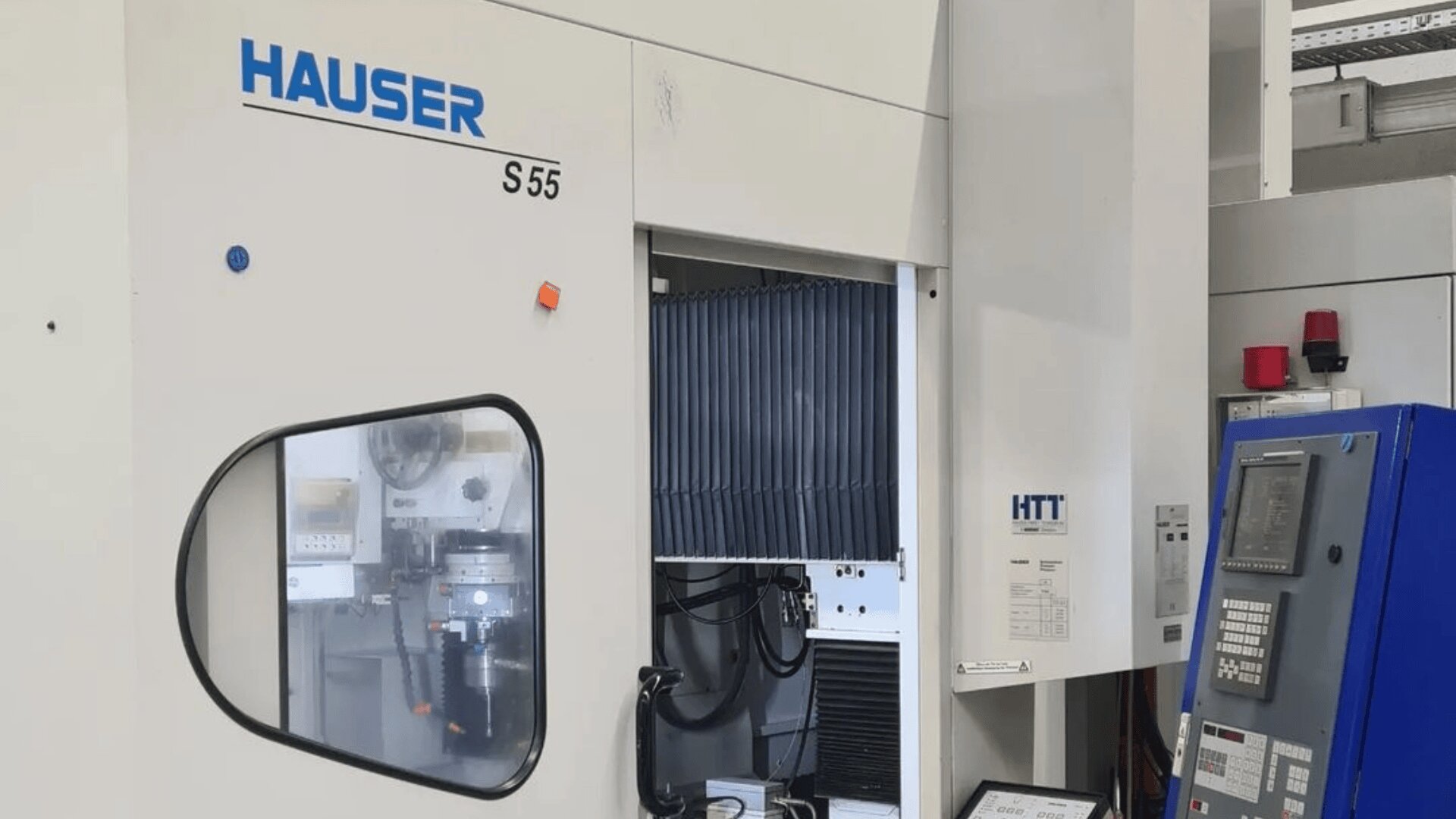 Eine Hauser S55 Industriemaschine mit Bedienfeldern und Displays. Ein offenes Fach zeigt interne Komponenten und Verkabelung. Die Maschine ist weiß mit blauem Text und ist von verschiedenen Geräten umgeben.