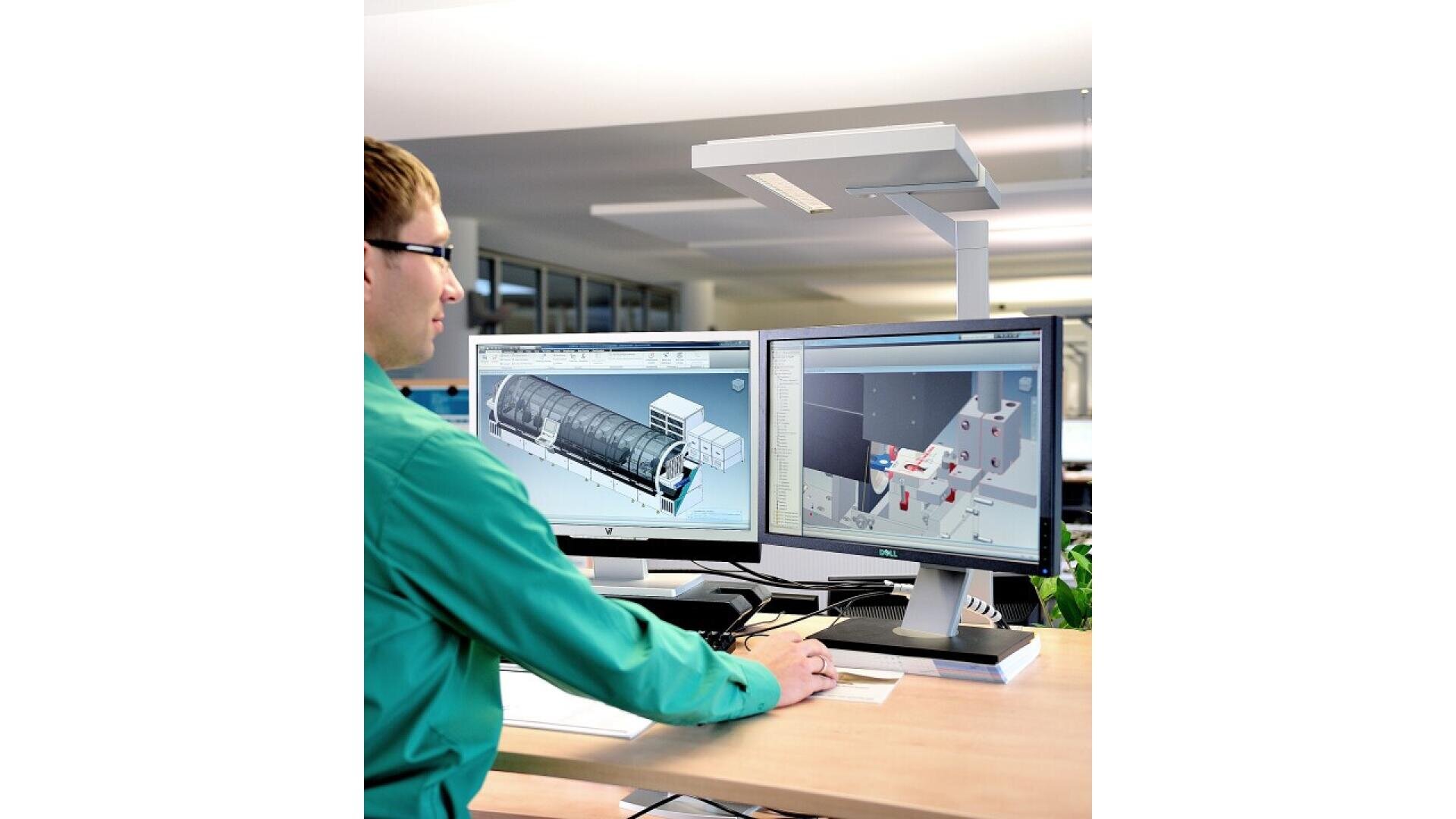A person in a green shirt sits at a desk using a computer with two monitors displaying 3D engineering or architectural designs in a modern, bright office environment.