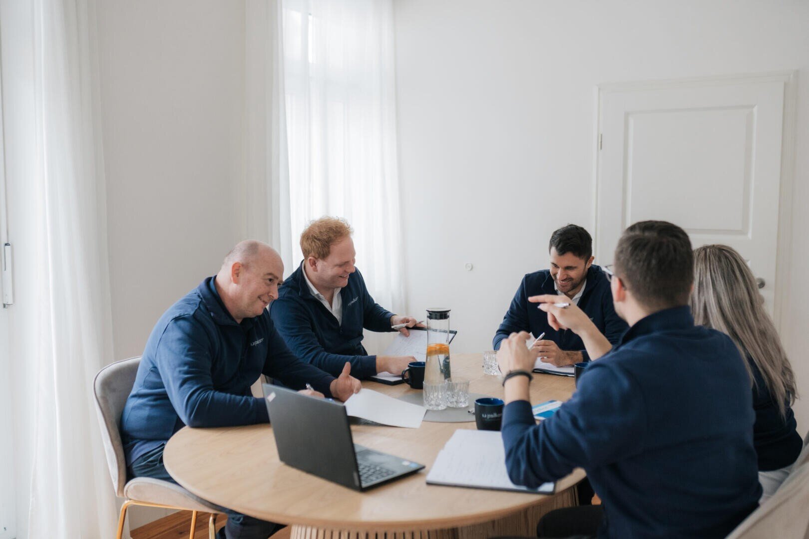 Fünf Personen sitzen um einen runden Tisch in einem hellen Raum und führen eine Besprechung durch. Laptops, Papiere und Getränke stehen auf dem Tisch, und die Gruppe scheint eifrig zu diskutieren.