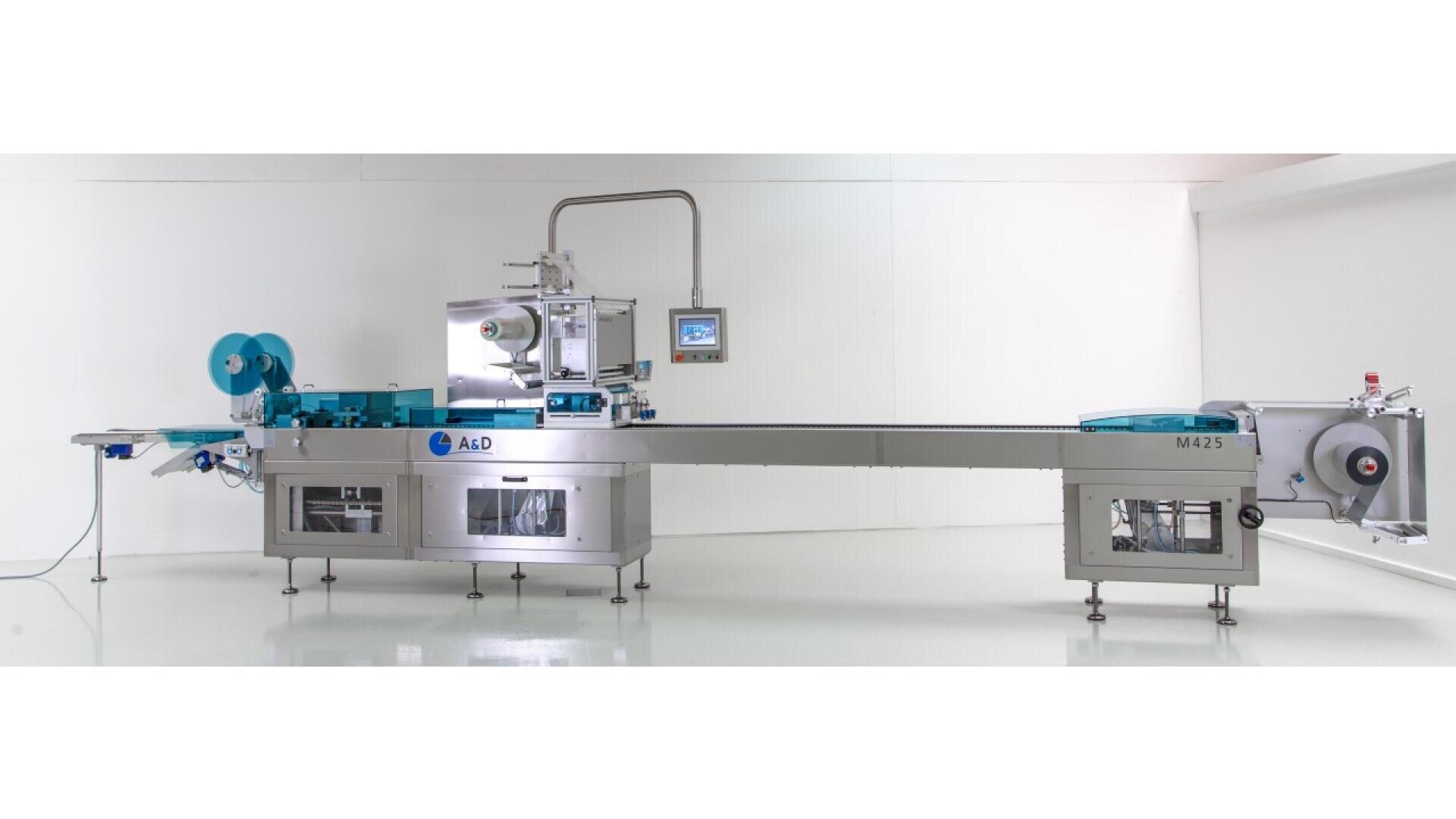 A large, industrial horizontal flow wrapping machine with blue and silver components, control panels, conveyor belts, and rolls of packaging film, set in a clean, white room.