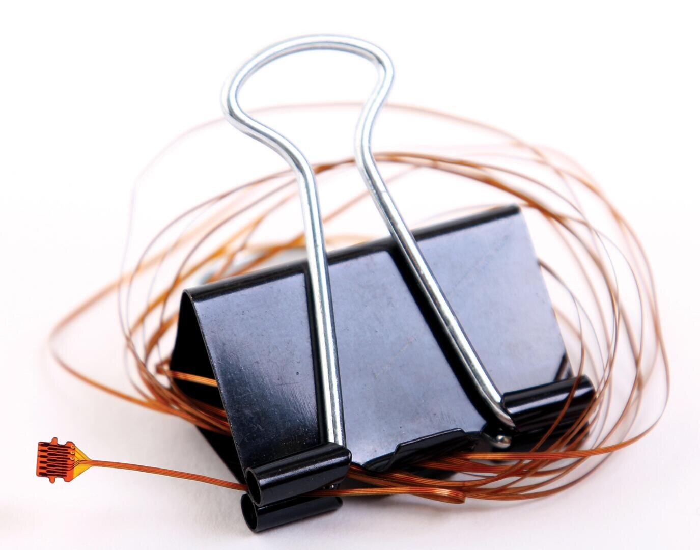 A black binder clip with a thin, coiled copper wire wrapped around it, and a small electronic sensor attached to the end of the wire, all set against a white background.