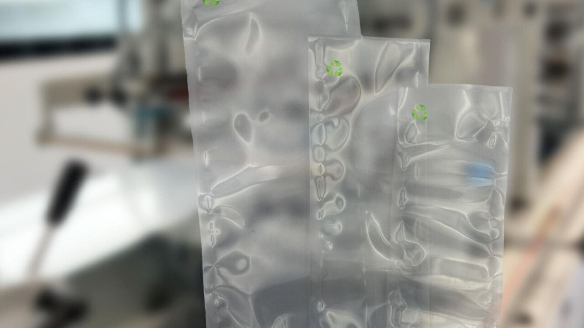 Three transparent inflatable air cushions of varying sizes are shown standing upright, used for packaging. Each has a small green recycling symbol. The background appears to be a blurred industrial or manufacturing setting.