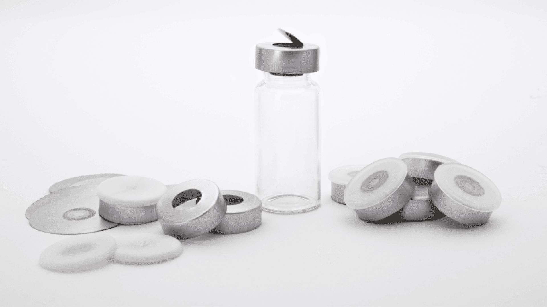 A clear glass vial with a partially opened metal cap is surrounded by several loose metal and plastic vial caps and seals, all set against a white background.