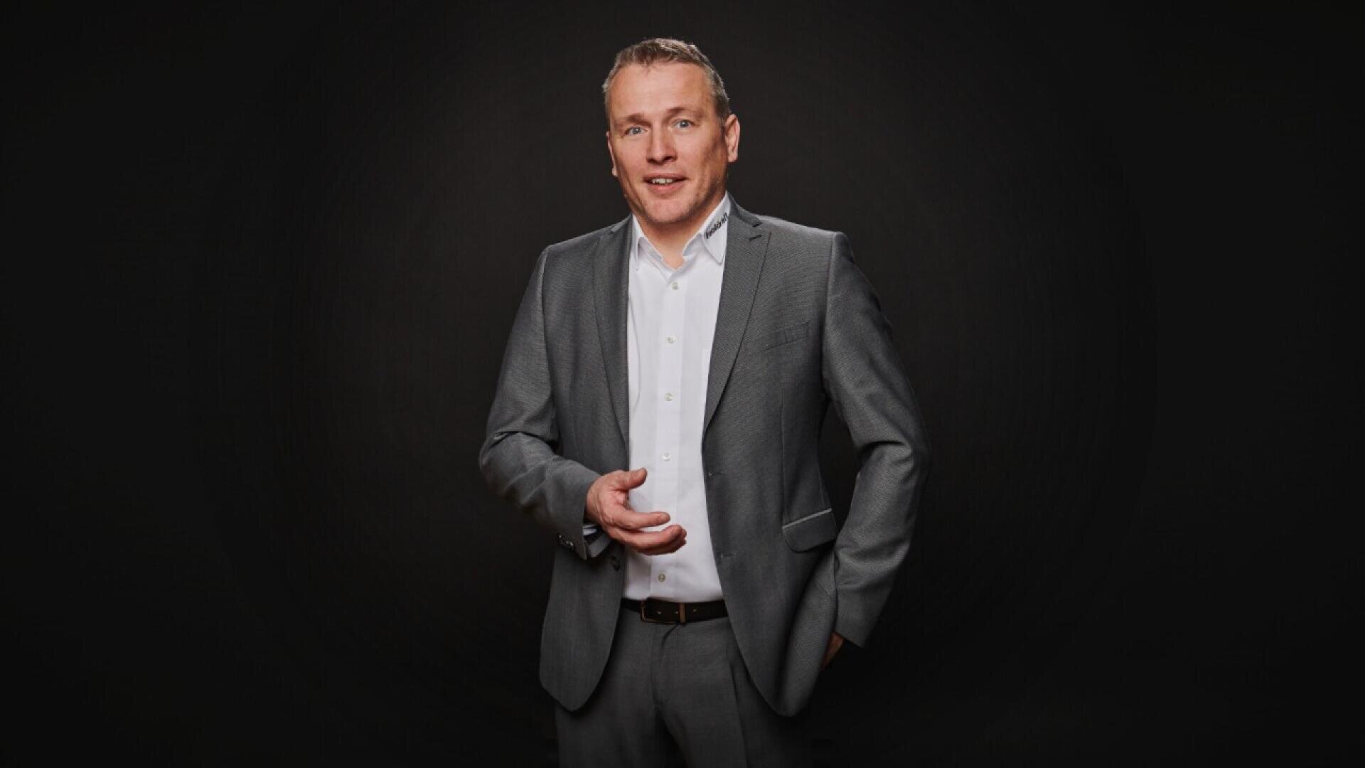 A man in a gray suit and white dress shirt stands against a dark background, facing the camera with one hand in his pocket and the other gesturing slightly, smiling.