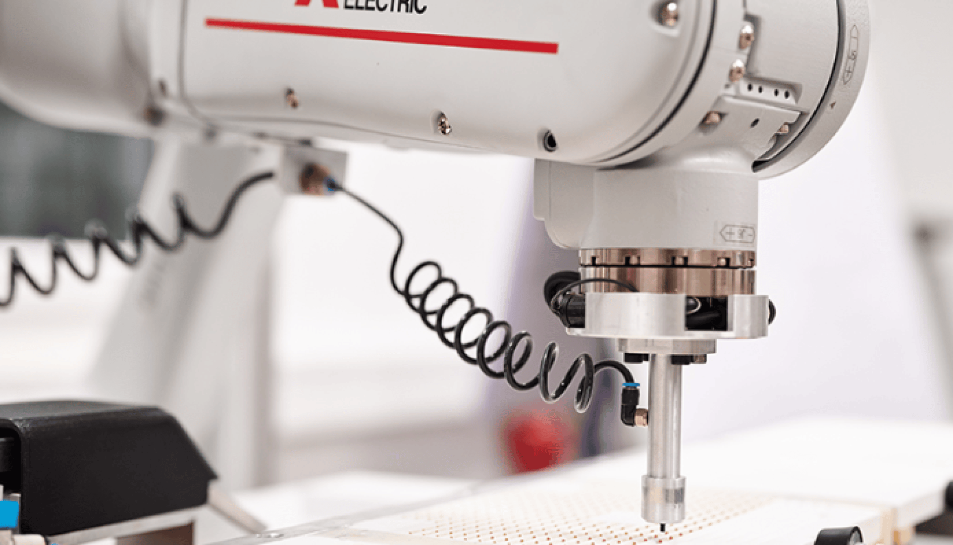 Close-up of an industrial robot arm with a metal tool attachment showing the word "ELECTRIC" and a coiled cable, working over a perforated surface in a clean, modern environment.