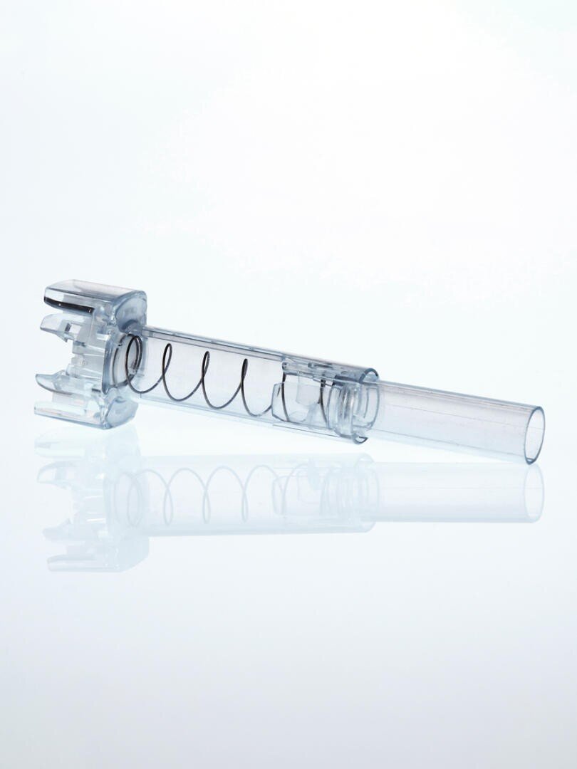 A transparent plastic medical device with a cylindrical shape and a visible metal spring inside, resting on a reflective white surface.