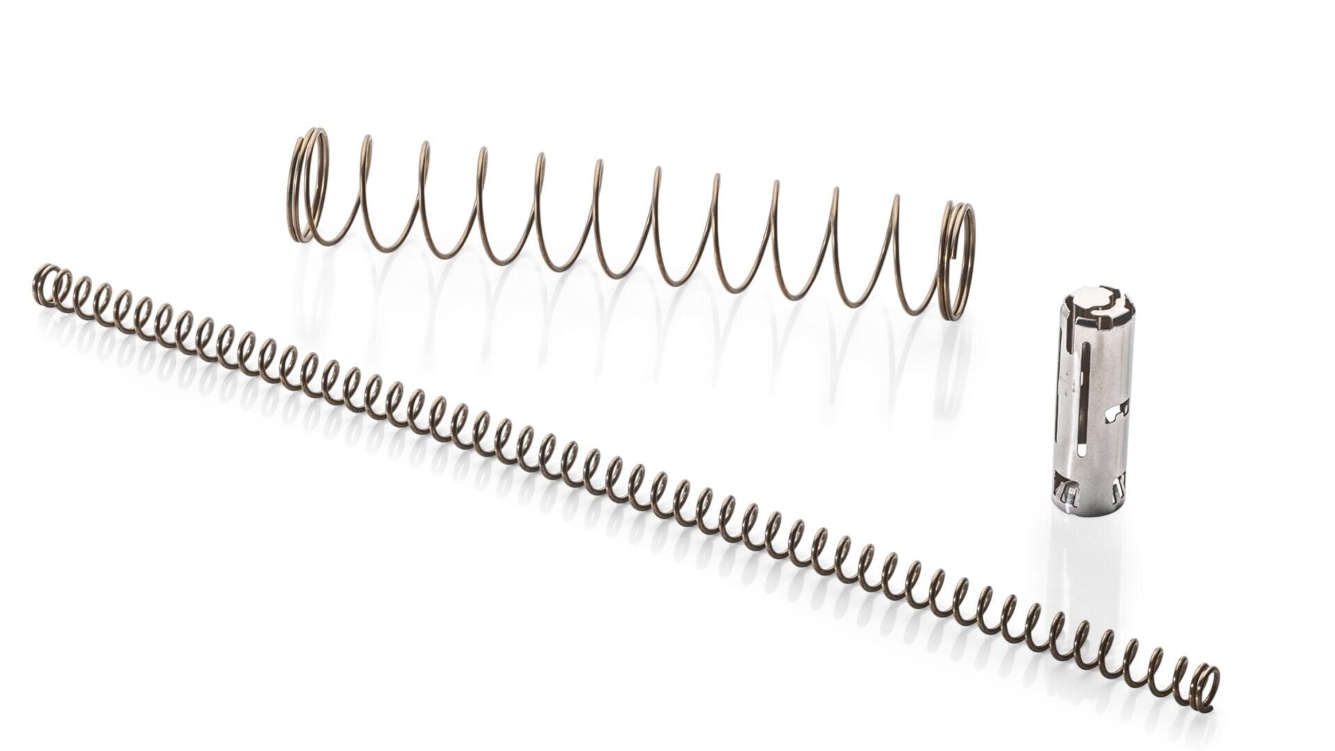 Three mechanical parts on a white background: a large coiled metal spring, a long thin tightly wound spring, and a small cylindrical metal component with slots and cutouts.