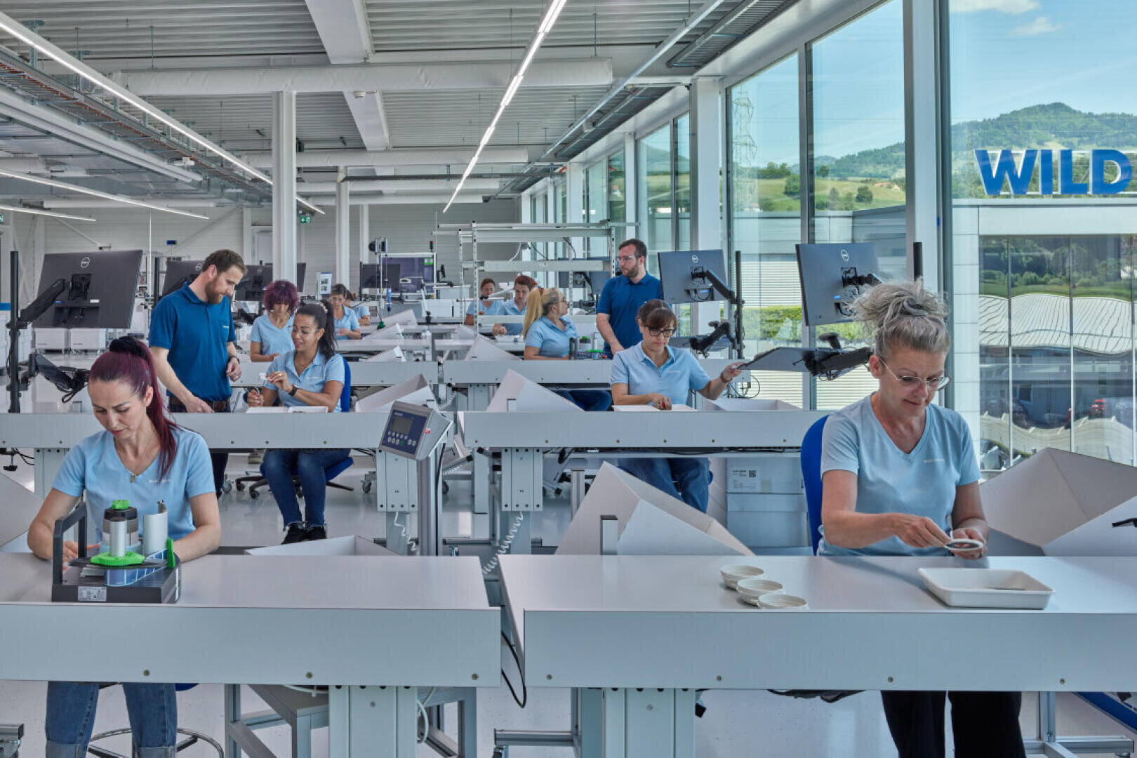 Workers, mostly women, sit at white desks in a bright, modern factory or laboratory. They are focused on assembling or inspecting small parts. Large windows show a scenic view of hills outside.