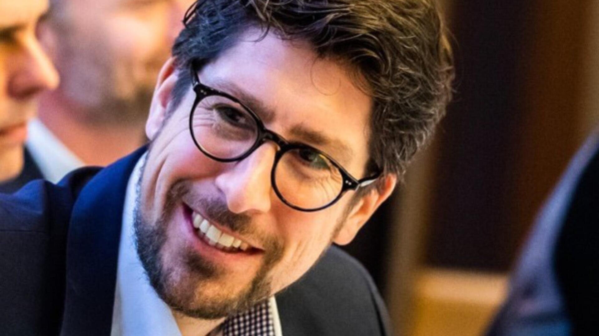 A man with dark hair, glasses, and a beard is smiling while wearing a suit and checkered shirt, sitting indoors with blurred people in the background.