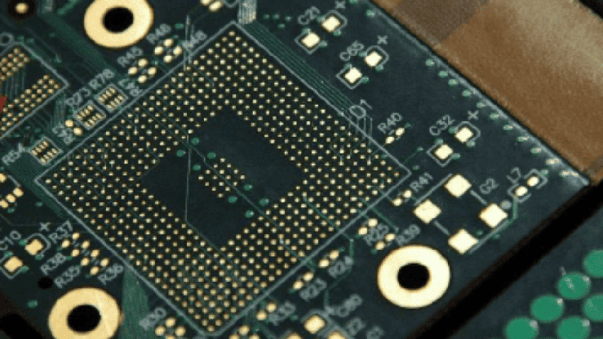 Nahaufnahme einer grünen Leiterplatte (PCB) mit zahlreichen goldenen Kontaktpunkten, runden Anschlüssen und verschiedenen beschrifteten elektronischen Komponenten.