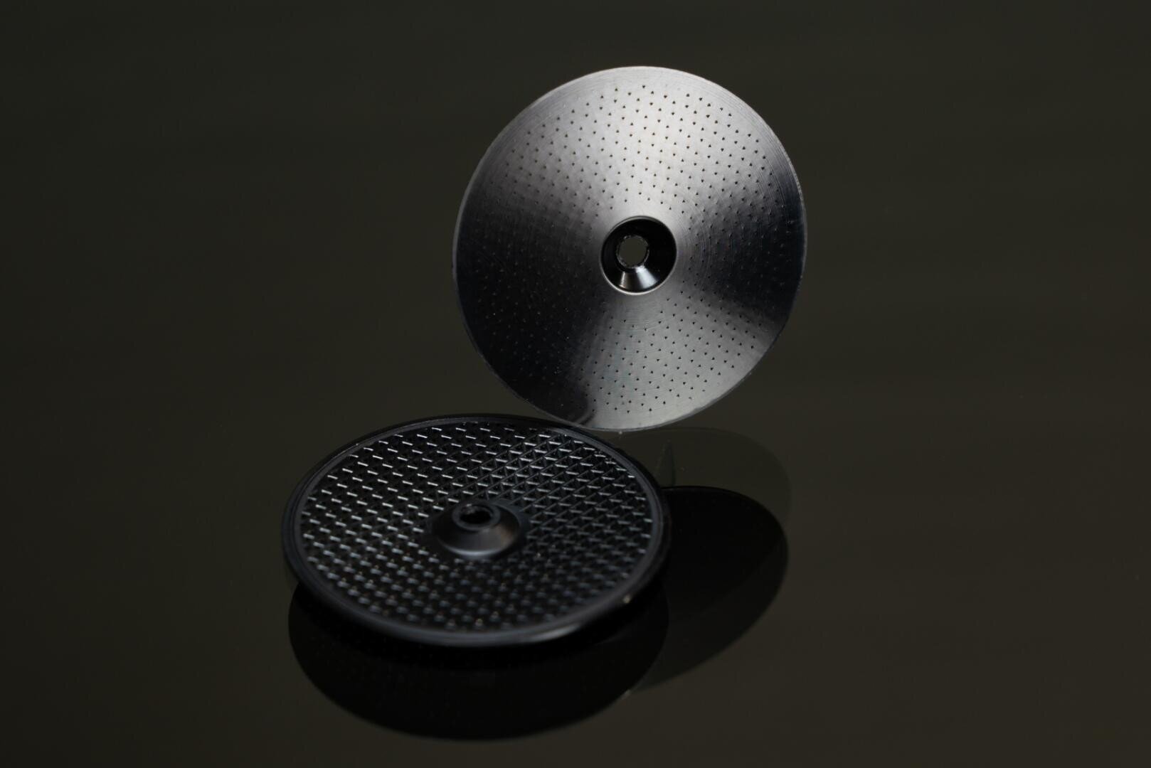 Zwei runde Metallscheiben mit kleinen Löchern; eine Scheibe steht aufrecht, während die andere flach liegt. Beide haben ein Loch in der Mitte und sind vor einer dunklen, reflektierenden Oberfläche ausgestellt.