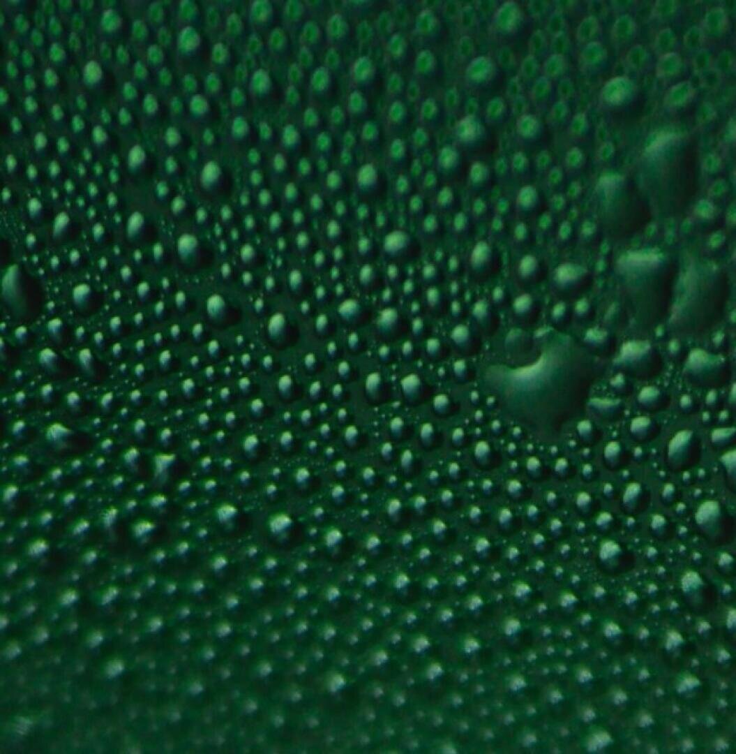 Nahaufnahme zahlreicher kleiner Wassertropfen auf einer grünen Oberfläche, die ein strukturiertes und gepunktetes Erscheinungsbild mit Tropfen unterschiedlicher Größe im gesamten Bild erzeugen.