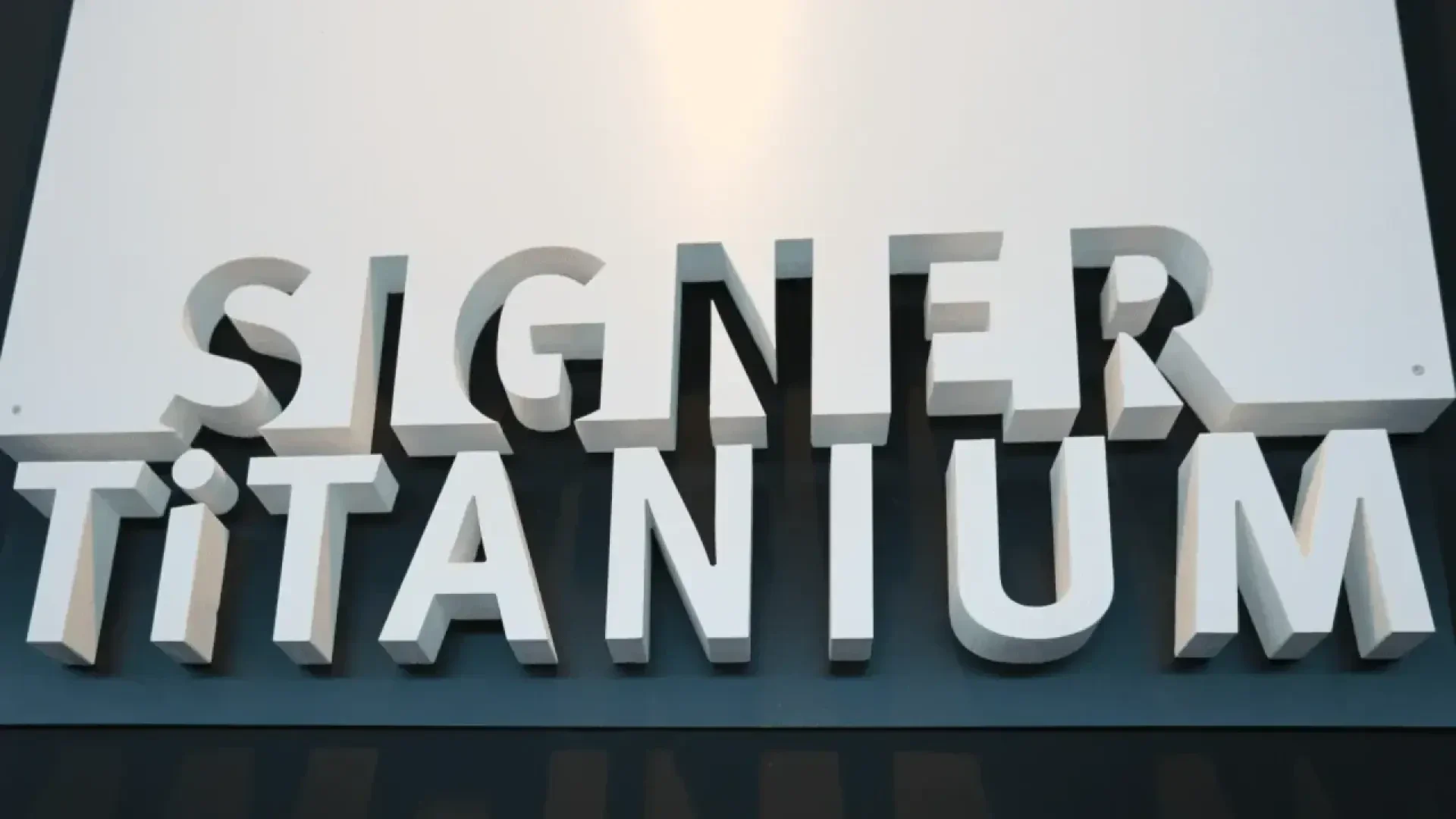 3D block letters spelling SIGNER TITANIUM are mounted on a wall, with light and shadow creating a bold, prominent effect.