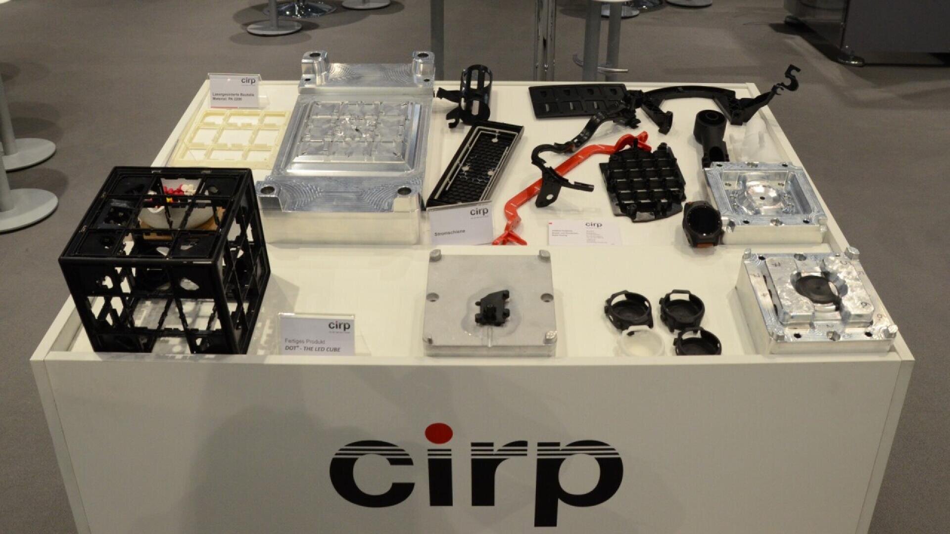 A white display table with various industrial parts and molds, including metal and plastic components. The table has the word cirp on its front, and several items are labeled with small cards.