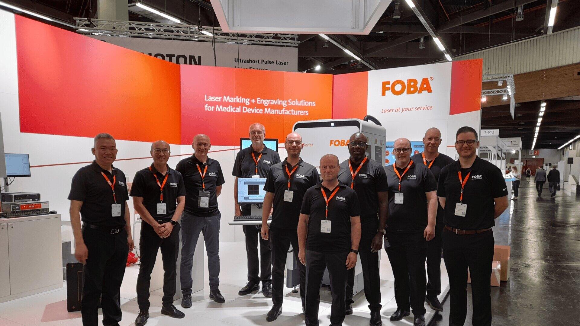 Eine Gruppe von elf Männern in passenden schwarzen Hemden mit orangefarbenen Schlüsselbändern steht lächelnd am Messestand von FOBA, einem Unternehmen, das sich auf Lasermarkierungs- und Gravurlösungen für Hersteller medizinischer Geräte spezialisiert hat.