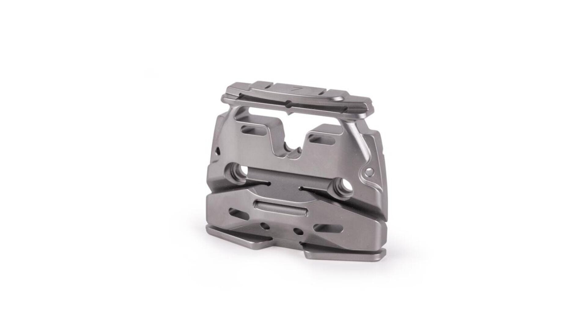 Ein metallisches, kantiges mechanisches Teil mit mehreren Schlitzen, Löchern und Rillen ist vor einem weißen Hintergrund abgebildet. Das Teil scheint präzisionsbearbeitet zu sein und hat eine matte silberne Oberfläche.