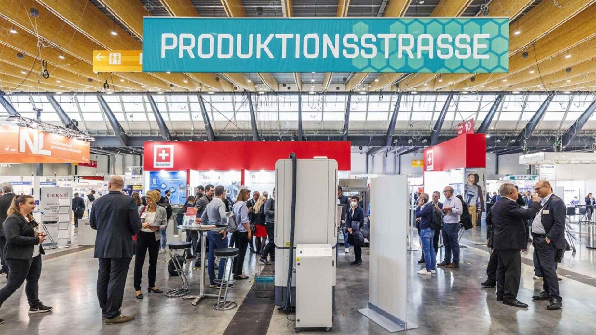 A busy exhibition hall with people standing and talking under a large sign reading PRODUKTIONSSTRASSE. Various booths, banners, and displays are visible in the background.