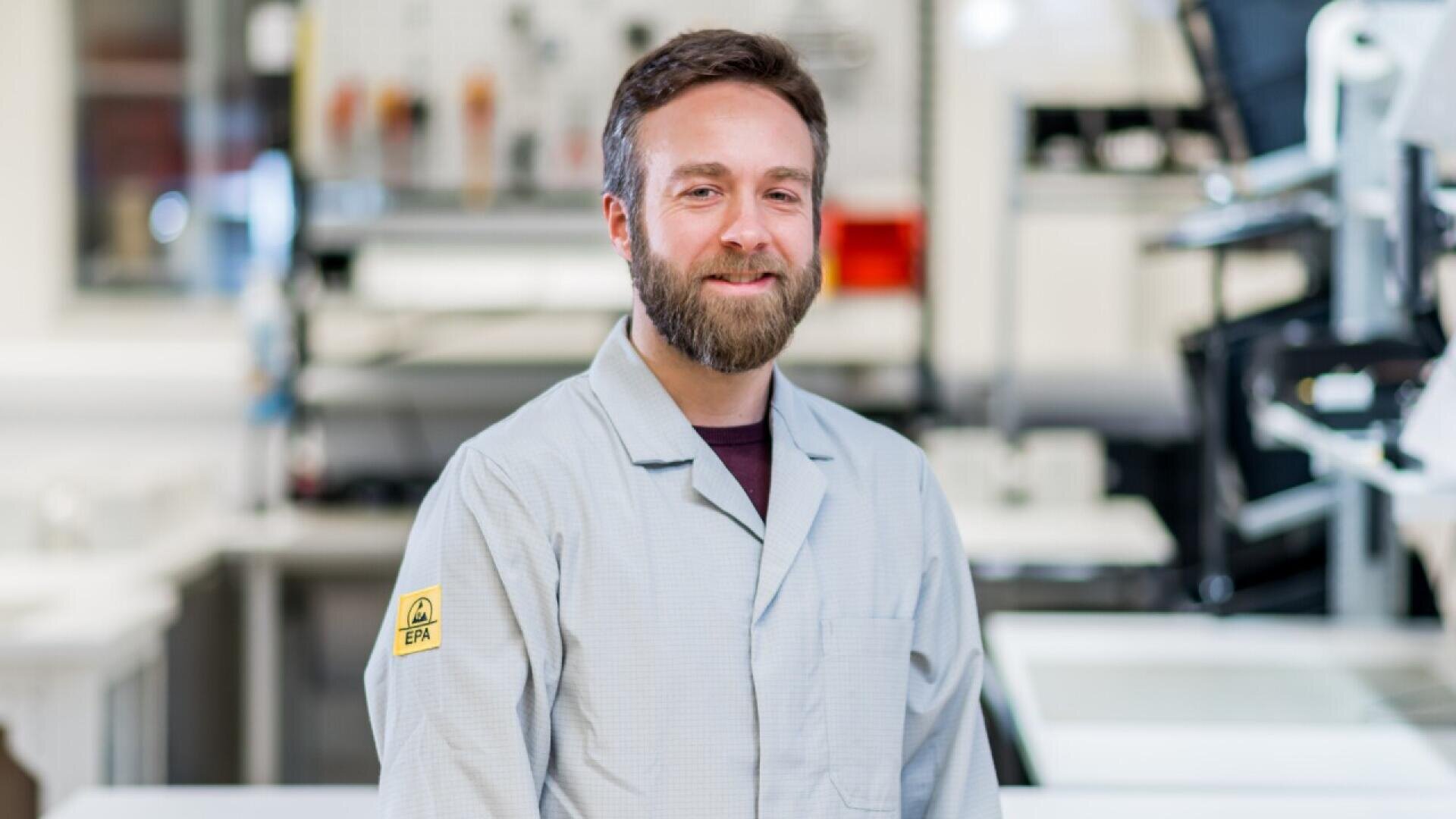 A man with a beard, wearing a light gray lab coat, stands smiling in a bright laboratory setting with scientific equipment in the background.