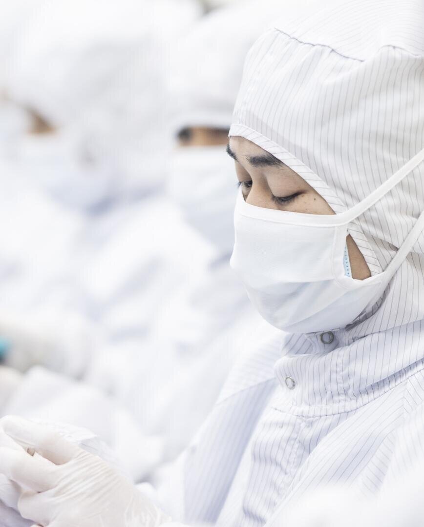 A person wearing a white protective suit, mask and gloves works in a cleanroom environment, while other people in similar clothing can be seen blurred in the background.