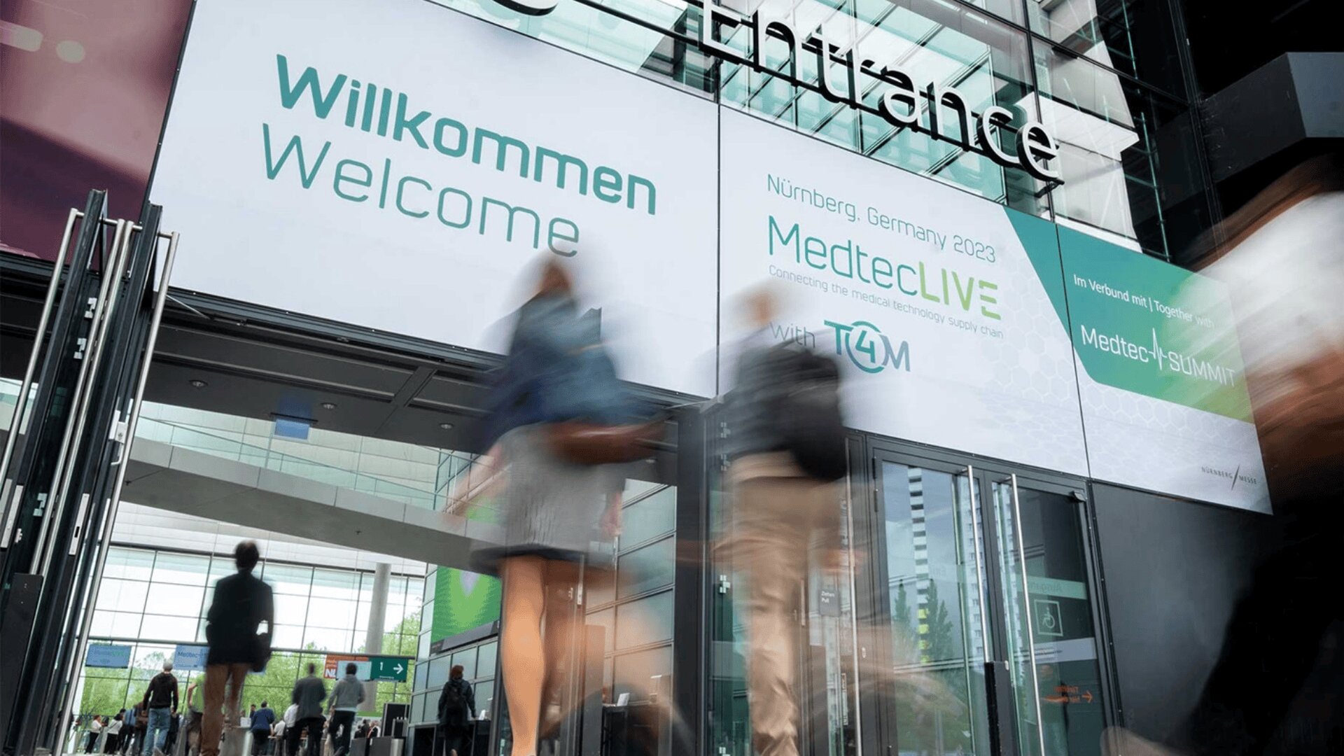 Die Menschen betreten ein modernes Glasgebäude mit großen Schildern über dem Eingang, auf denen "Willkommen" und "MedtecLIVE Nürnberg Germany 2023" steht. Die Szene ist lebhaft und die Bewegungen der Teilnehmer verschwimmen.