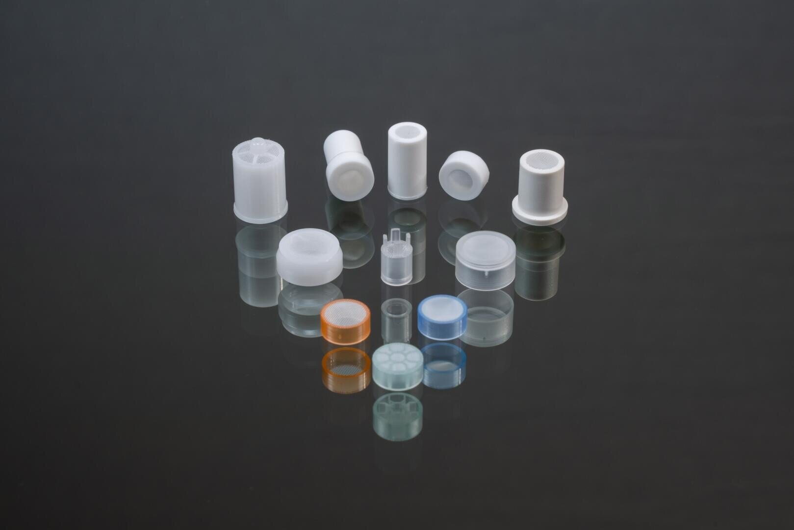 Eine Vielzahl kleiner, zylindrischer Kunststoff- und Gummiteile, teils durchscheinend, teils farbig, sind auf einer schwarzen, reflektierenden Oberfläche übersichtlich angeordnet.
