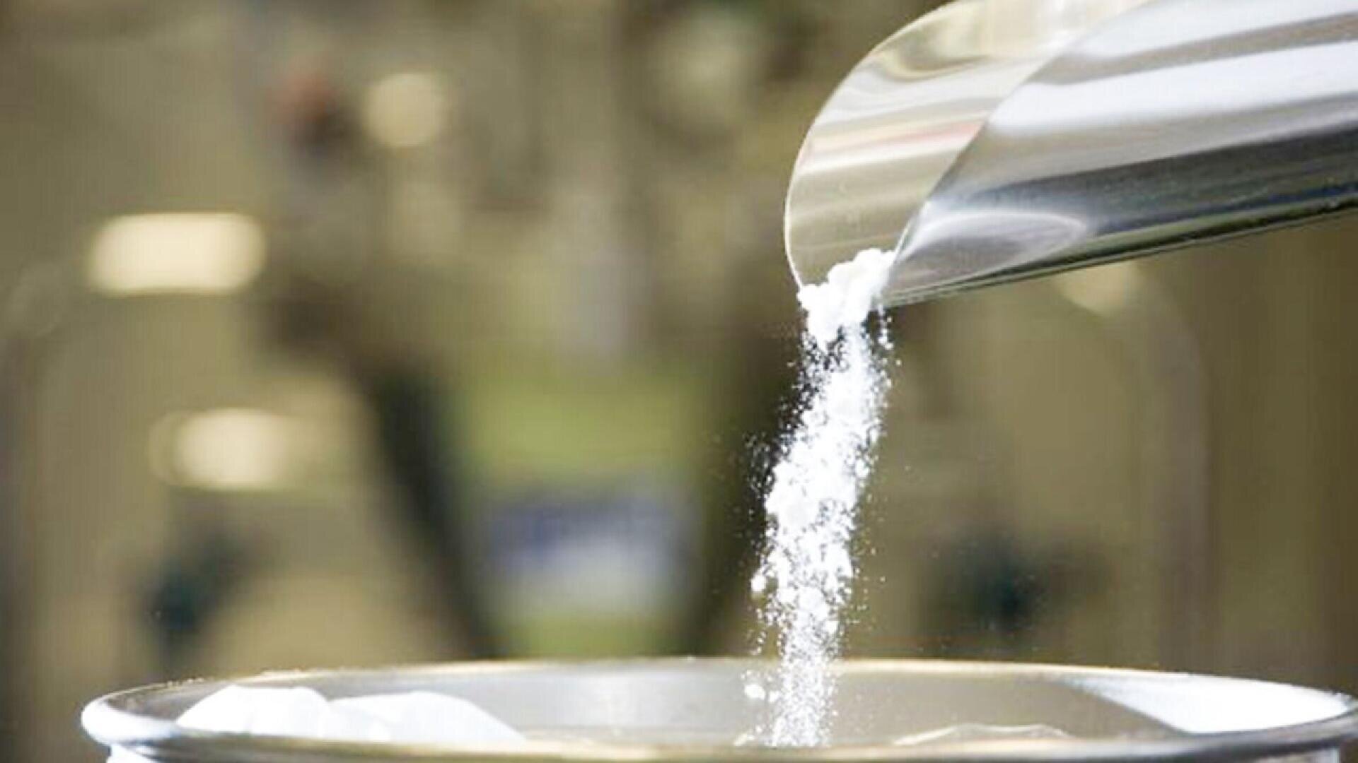A close-up of white powder being poured from a metal scoop into a metal container, with a blurred background.